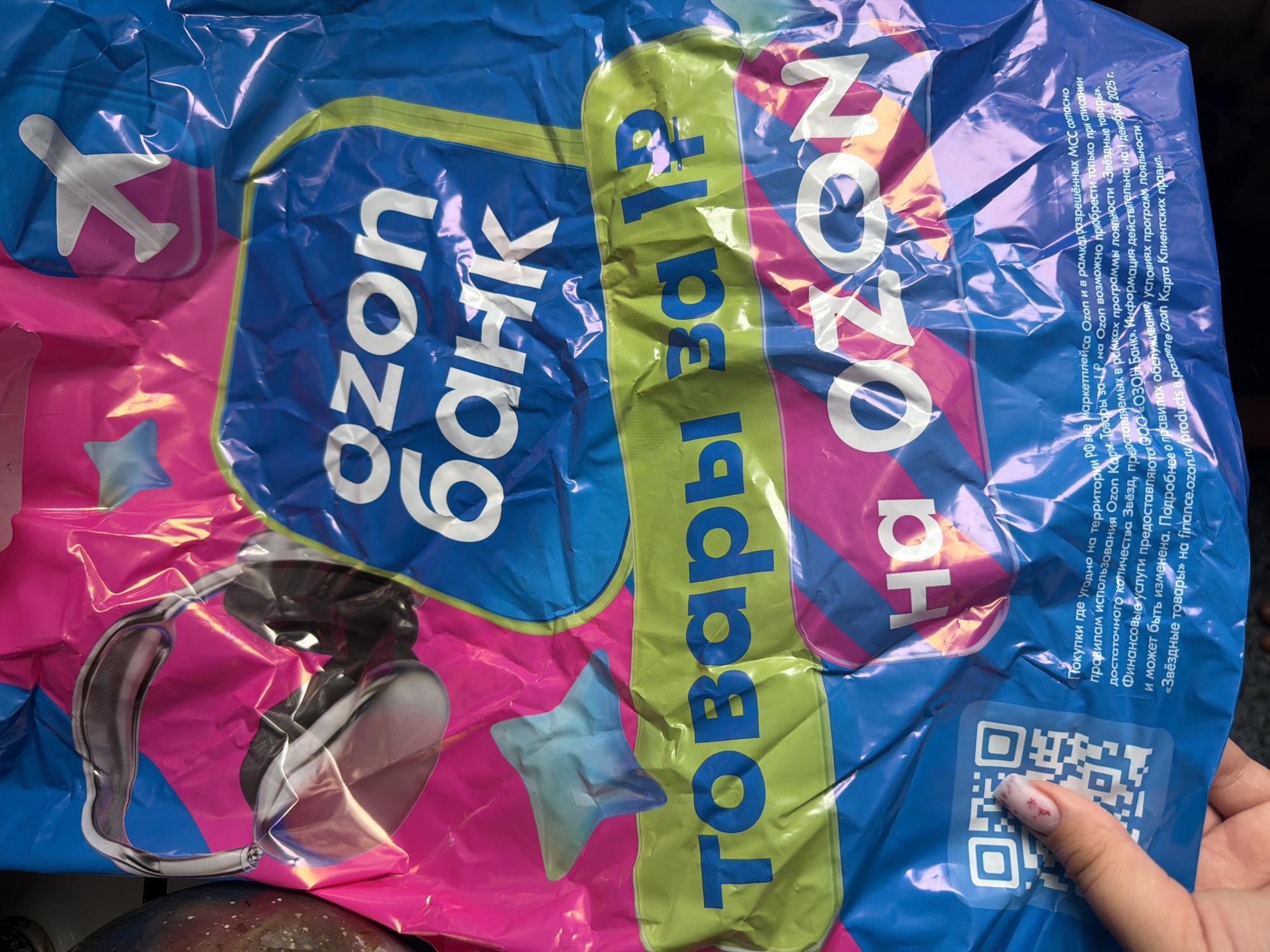 The image shows a close-up of a crumpled plastic bag, likely packaging for a product or service. The bag features bold, colorful branding with the text "OZON BANK" prominently displayed in white letters on a blue background. Below this, the words "Товары за 1р" (Goods for 1 ruble) are written in blue on a green band. Another large white text reads "OZON". There are also graphics of headphones, an airplane, and a star. A QR code is visible on the lower left side, partially obscured by a person's finger with a pink and red patterned fingernail. Small text in Russian is printed along the bottom of the bag, providing details about purchases and terms and conditions. The bag appears to be made of a glossy, reflective material that is creased and distorted, suggesting it has been handled or is being stored. The background is out of focus, but appears to be a dark, possibly indoor environment. No specific location in the city of Chaikovsky, Russia is indicated in the image.