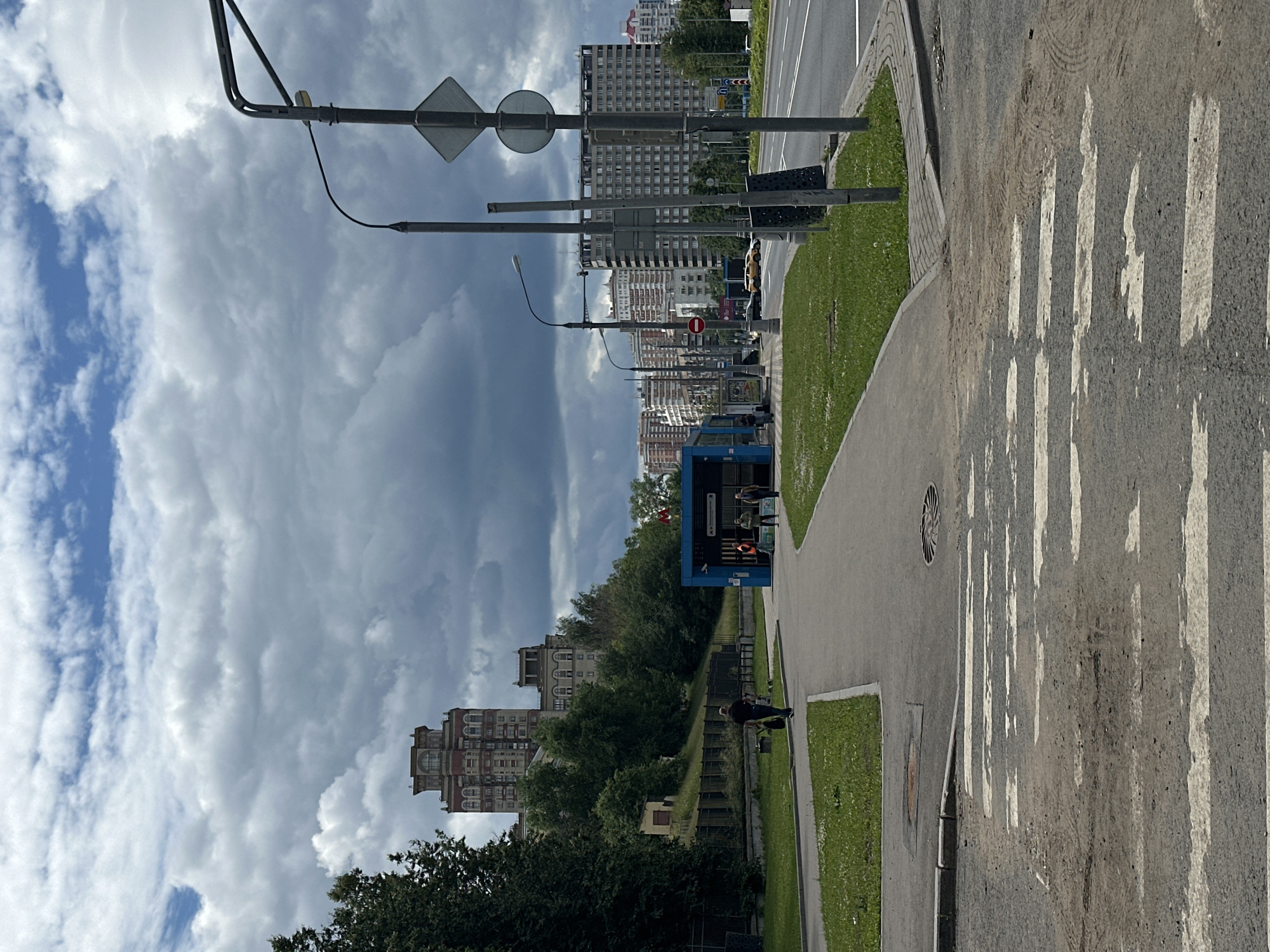 The image shows a pedestrian crossing in front of a blue metro station entrance in Moscow, Russia. The sky is overcast with dramatic clouds. In the background, there are several multi-story apartment buildings and trees. A man is walking away from the camera, pulling a cart, on the sidewalk leading to the metro station. Two other people are standing near the entrance of the station. The street adjacent to the sidewalk has cars driving on it, including a yellow car. The pedestrian crossing is in the foreground, showing worn white stripes on asphalt.