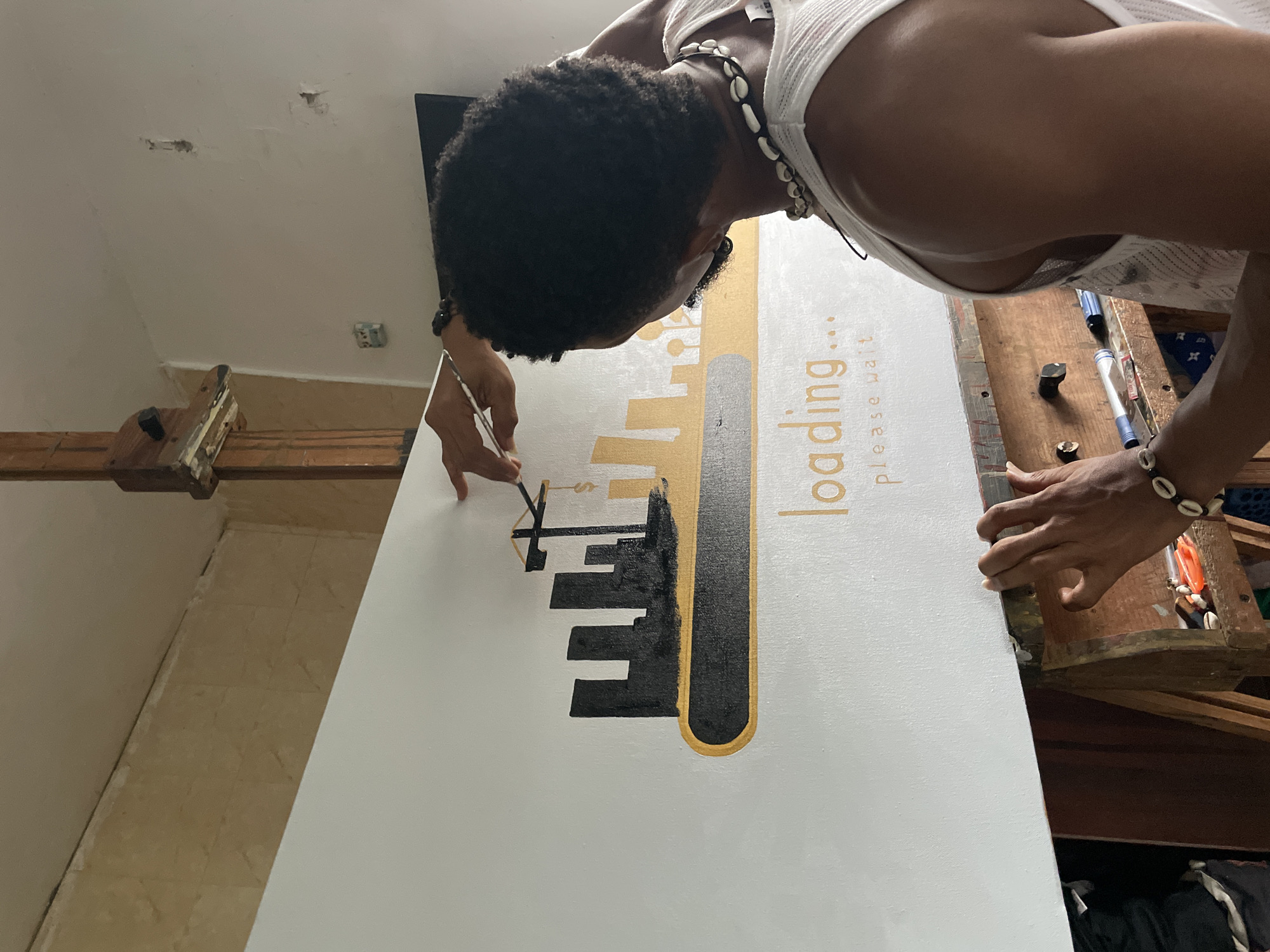 A person is painting a large canvas with a design that incorporates the word "loading..." and a skyline motif. The person, likely an artist, is wearing a white tank top and shell jewelry. They are using a fine brush to apply black paint to the canvas. The painting features gold accents. The environment appears to be an indoor studio or workspace. The location is Ado, Nigeria.