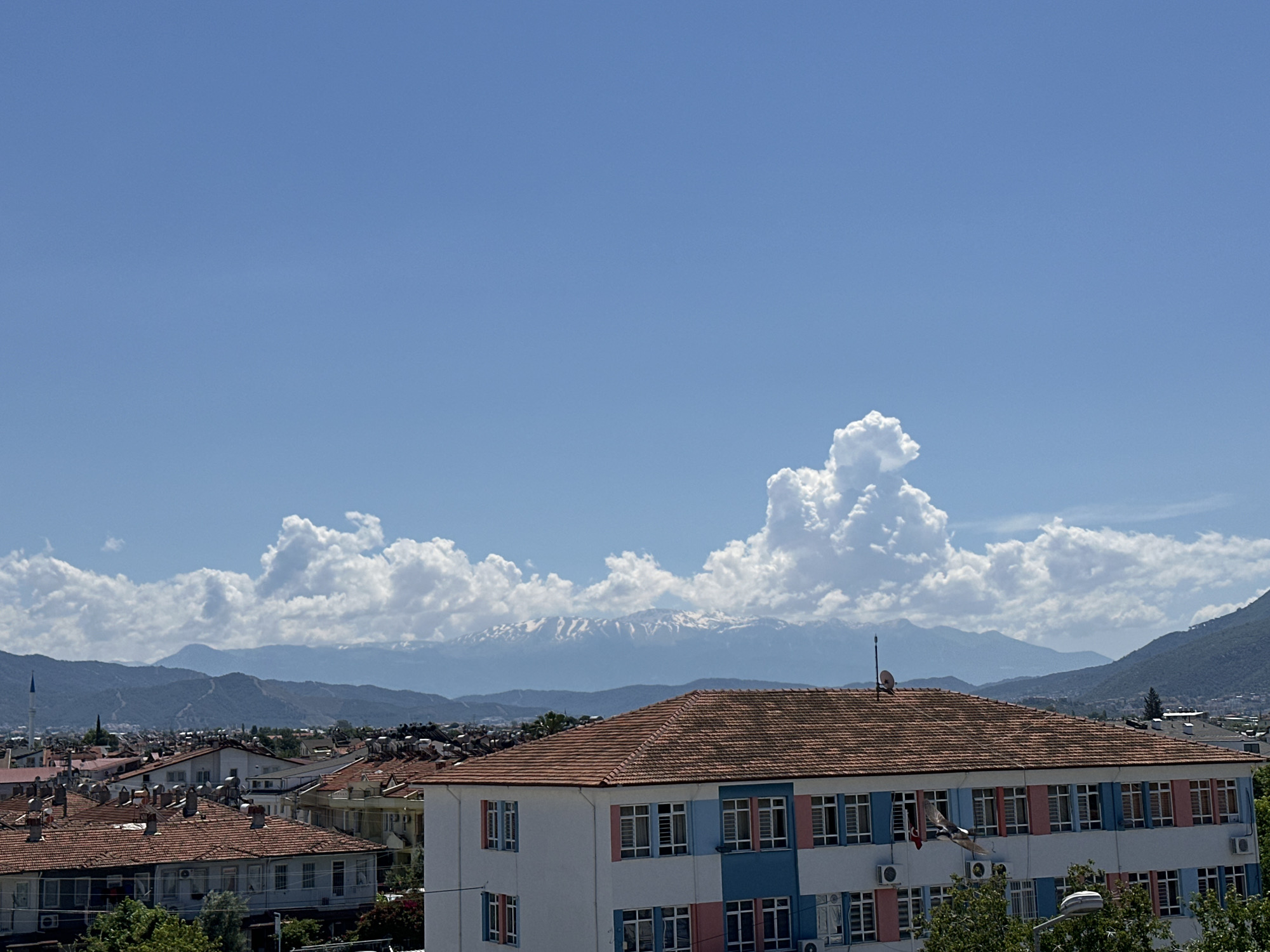 The image captures an elevated view of Fethiye, Turkey, under a blue sky with scattered cumulus clouds. A mountain range with visible snow caps sits in the middle ground. Below the mountains, a densely packed urban landscape is visible, comprised of buildings with red-tiled roofs. Dominating the foreground is a two-story building with a terracotta-colored tiled roof. The building's facade is white with blue and pink accents around the windows. Air conditioning units are mounted on the exterior walls. Mature trees partially obscure the lower portion of the building.