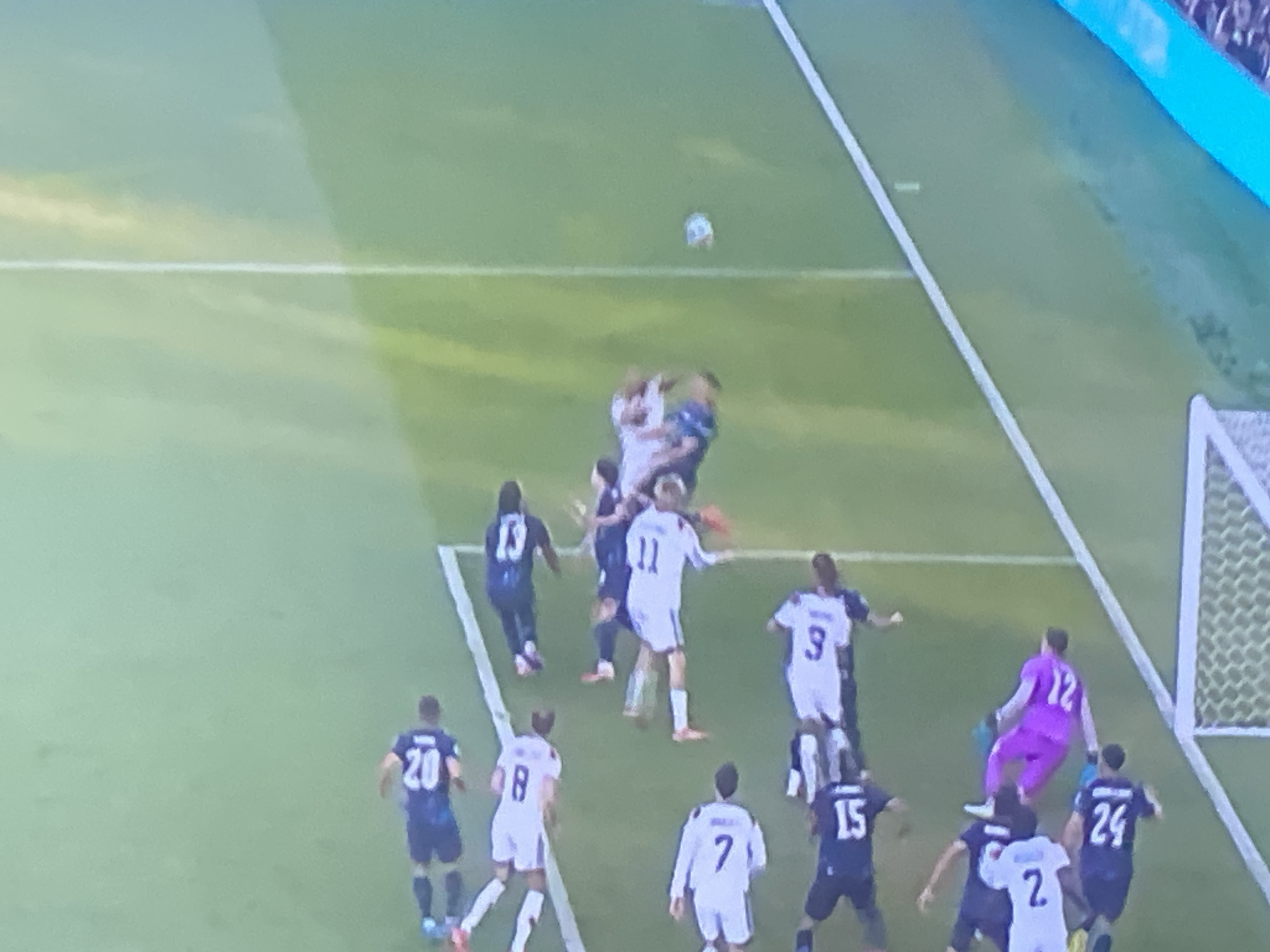 The image shows a soccer game in progress. The field is green with white lines. Several players are visible on the field, wearing blue and white uniforms. One player in a purple uniform, likely a goalkeeper, is near the goal. A soccer ball is in the air. The game appears to be at a pivotal moment, possibly a corner kick or a cross into the box. The image is of moderate resolution and is somewhat blurry, suggesting it was captured from a distance or on a lower-quality screen. There is no visible indication of the city of Zastavna, Ukraine, or any specific landmarks. The environment appears to be an outdoor sports stadium, with spectators visible in the background. The lighting suggests it is daytime, with some shadows cast on the field.