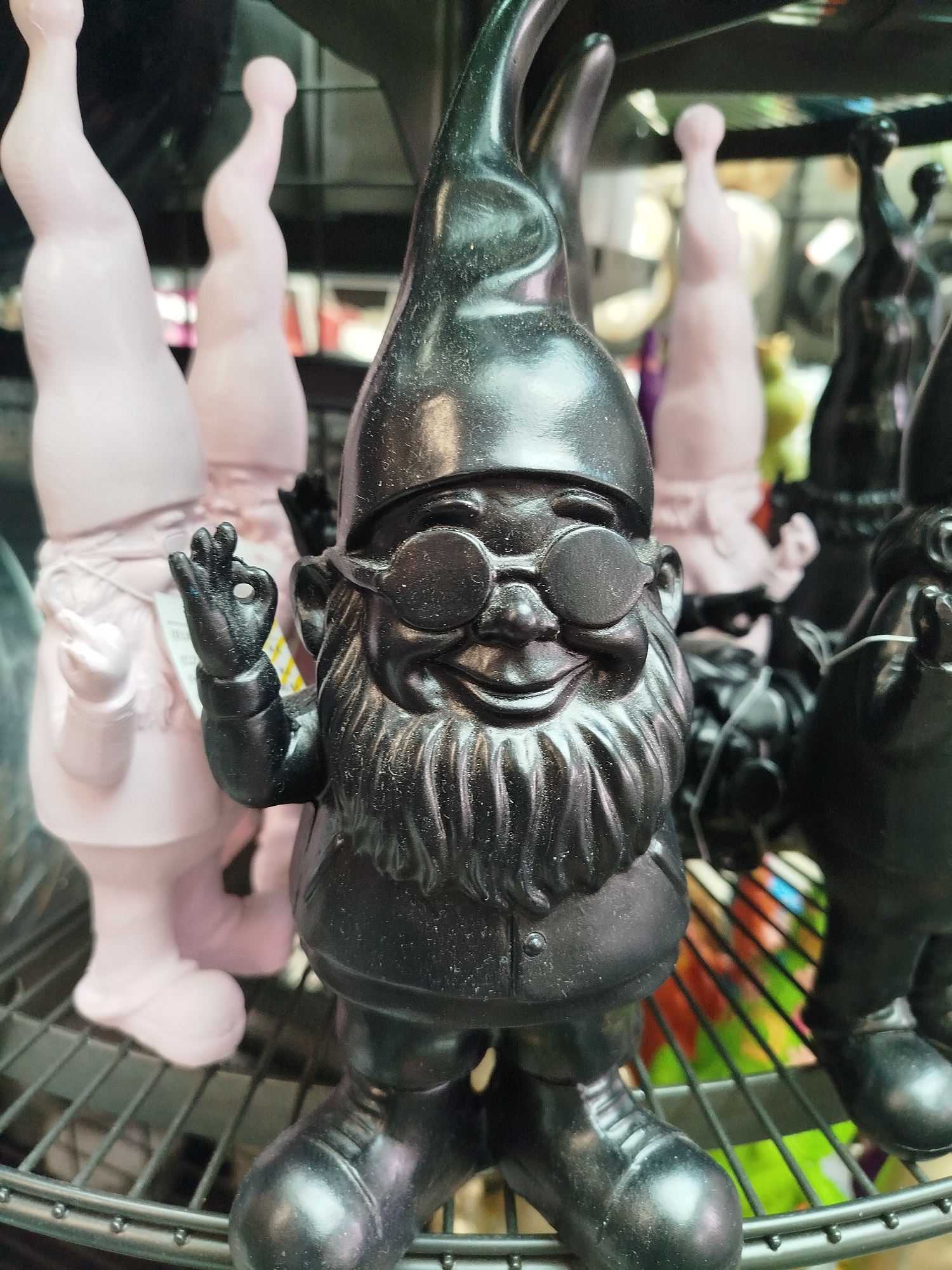 The image depicts a close-up view of several decorative garden gnomes displayed on a shelf. The gnome in the foreground is black and appears to be made of plastic or resin. It is wearing a pointed hat, round glasses, and has a prominent beard. Its right hand is raised, with the index finger and thumb forming a circle, a gesture often associated with the "OK" sign. Behind this gnome, several other gnomes are visible, some are light pink and others are black. These gnomes are also in various poses and appear to be part of a larger display. The background is blurred, but suggests an indoor retail environment, possibly a garden center or a novelty shop. There is no indication of the city of Offenbach am Main, Germany, or any specific location information within the image itself.