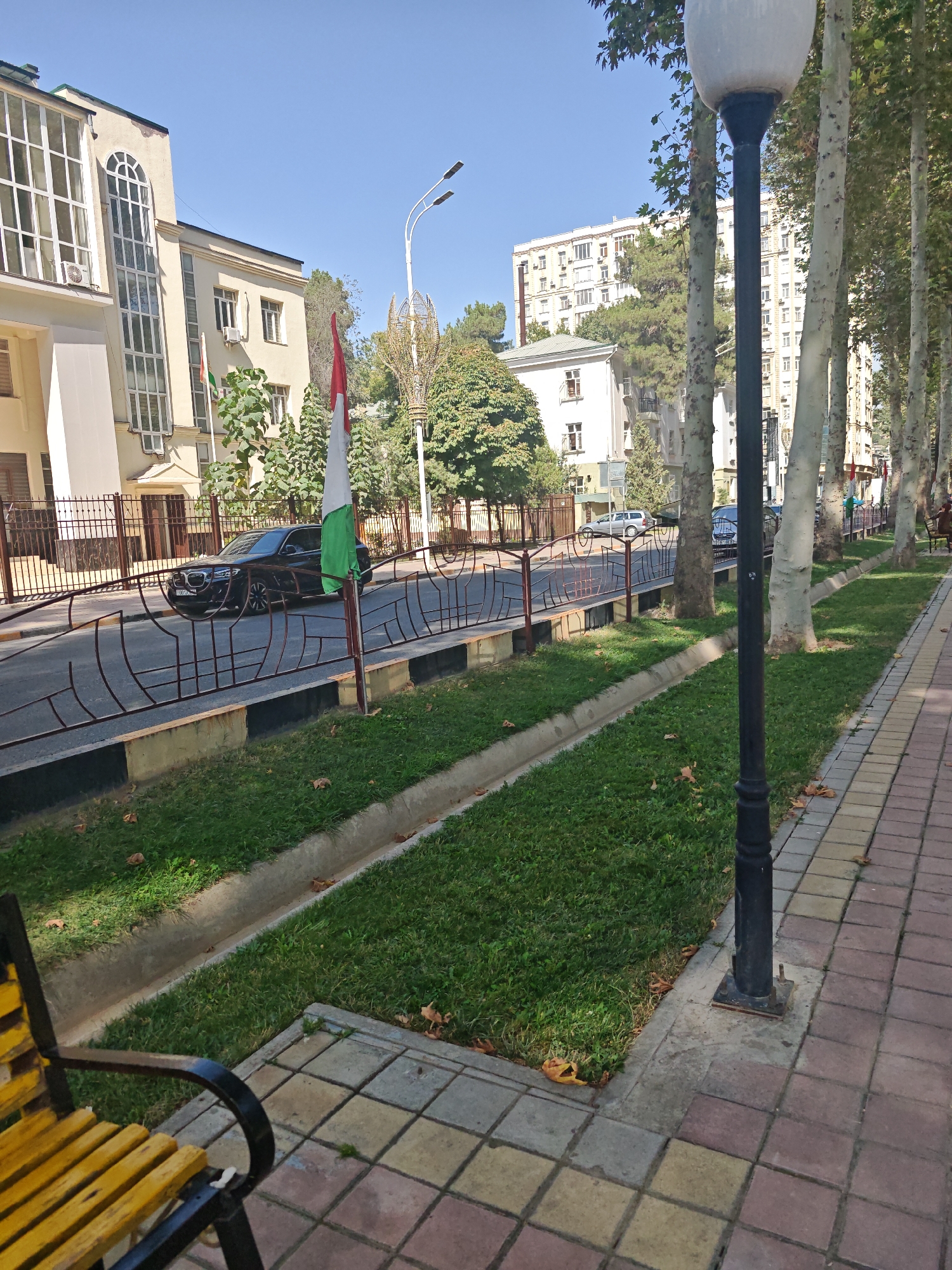 A sunny day in Unknown, Unknown, features a street lined with tall trees. On the left, a cream-colored building with large windows stands prominently. A black BMW is parked on the street, with a flag pole flying a tri-colored flag (red, white, and green) positioned in front of it. The street is bordered by a decorative metal fence. Adjacent to the street is a grassy area with a concrete drainage channel. The sidewalk on the right is paved with checkered tiles in shades of yellow, red, and grey. A black lamppost stands on the sidewalk, and a yellow and black park bench is visible on the far left. In the background, several other buildings of varying sizes and architectural styles are visible, including apartment buildings. The sky is clear and blue.