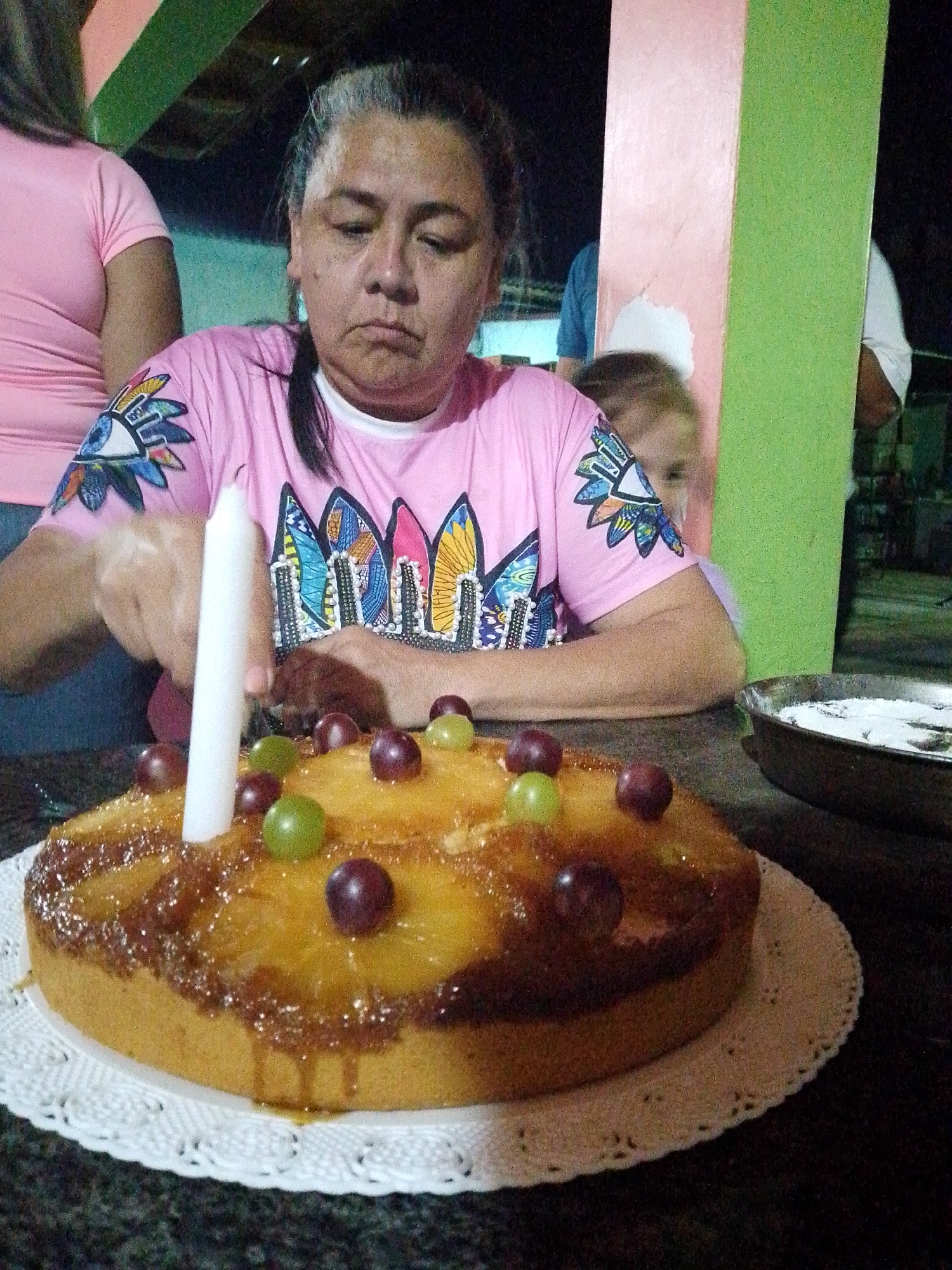 The image captures a woman with graying hair and a pink shirt adorned with colorful eye and wing motifs, sitting at a table indoors. In front of her is a round pineapple upside-down cake, decorated with green and purple grapes, and a single white candle. The cake sits on a white lace paper doily. To the right, there is a metal pan containing a white substance. The scene appears to be a birthday celebration. Behind the woman, glimpses of other people are visible. The location is Tinaquillo, Venezuela.