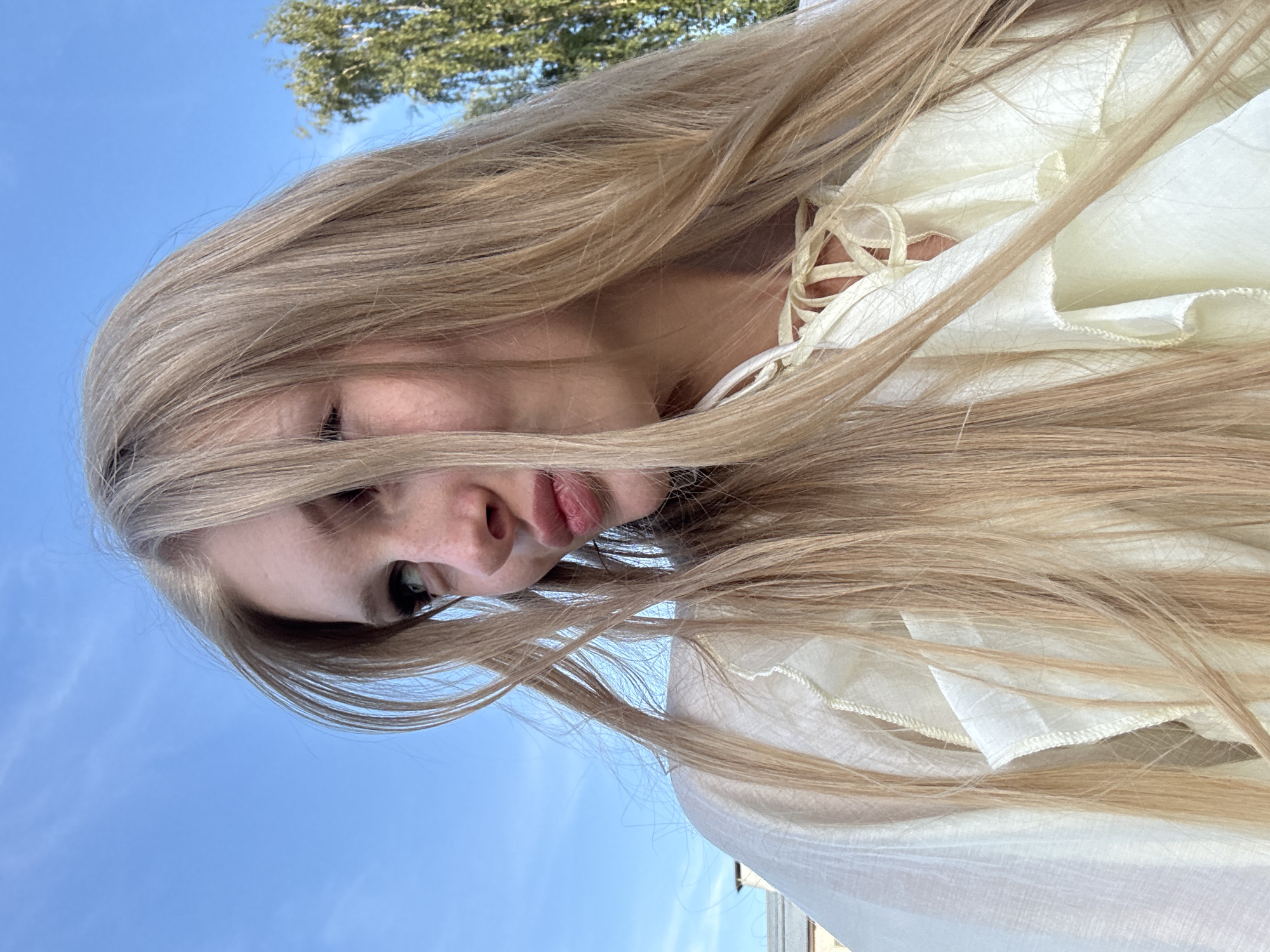 An outdoor, eye-level, medium shot shows a young Caucasian woman with long, blonde hair and fair skin, looking to her left and slightly upwards. Her lips are slightly parted, and she has freckles across her nose and cheeks. She is wearing a light-colored, ruffled garment with a lace-up neckline. The background consists of a bright blue sky with wispy clouds and the upper branches of a green tree on the right. The upper left of the image shows the corner of a light-colored building with a chimney. The image was taken on a sunny day.