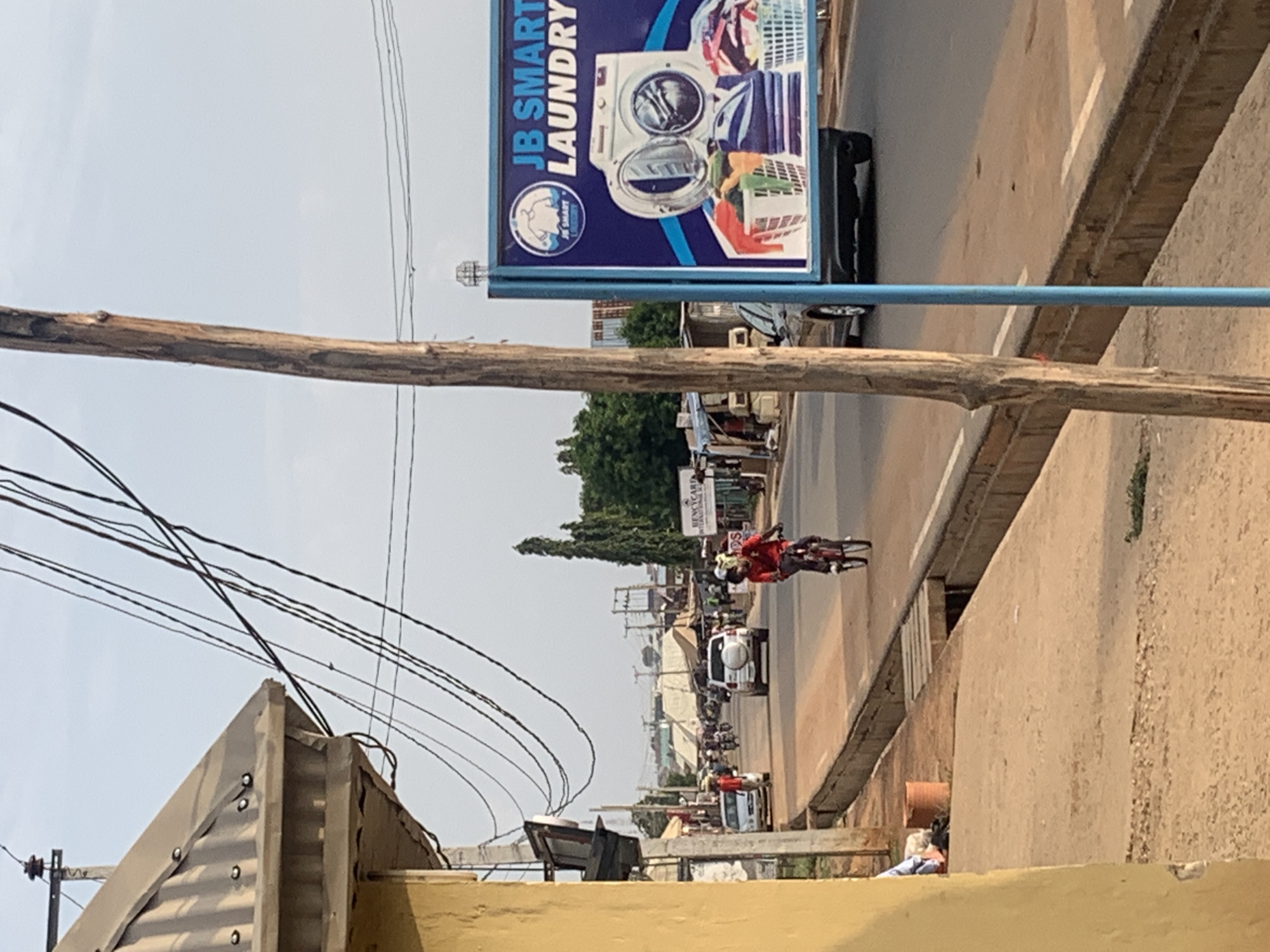 The image shows a street scene in Sabojibi, Nigeria. A utility pole and overhead wires are visible in the foreground. A "JB Smart Laundry" sign is on the left side of the street. Several vehicles, including cars and motorcycles, are on the road. A cyclist is riding towards the camera. Buildings and vegetation are visible in the background.