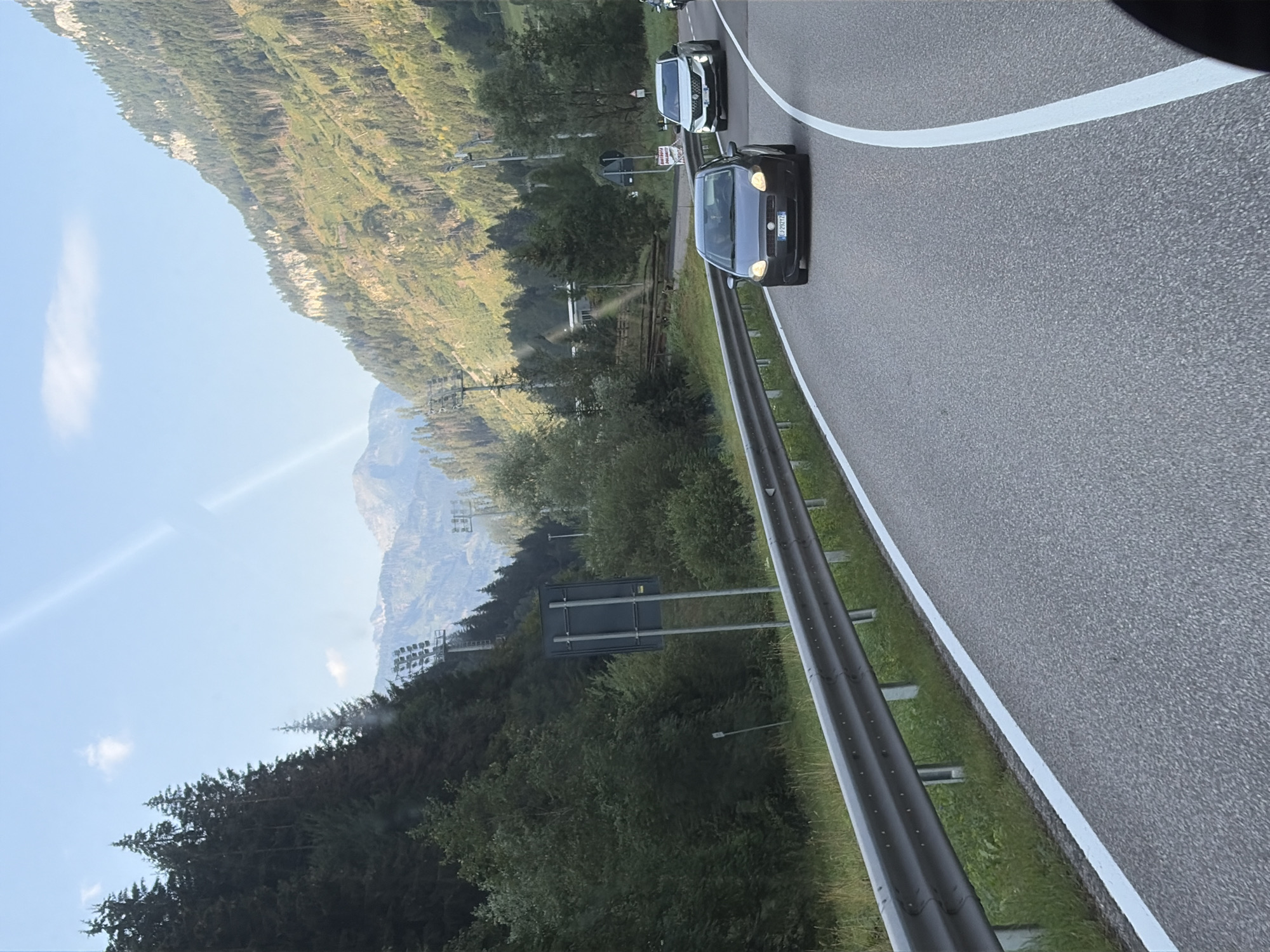 The image depicts a scenic mountain road in Predazzo, Italy, likely captured from within a vehicle. In the foreground, a dark-colored Fiat car is traveling on the right side of the asphalt road, which is marked with white lines. A metal guardrail runs along the left side of the road, separating it from a grassy embankment and a dense forest.
The surrounding environment is characterized by a steep, wooded mountainside on the left and a sun-drenched, rocky mountain slope on the right. The sky is clear blue with a few wispy clouds. In the distance, a series of pylons, possibly related to ski lifts or power lines, are visible ascending the mountainside. The lighting suggests it is daytime, with the sun casting strong shadows and illuminating the mountainous terrain. The overall impression is one of a picturesque alpine landscape.