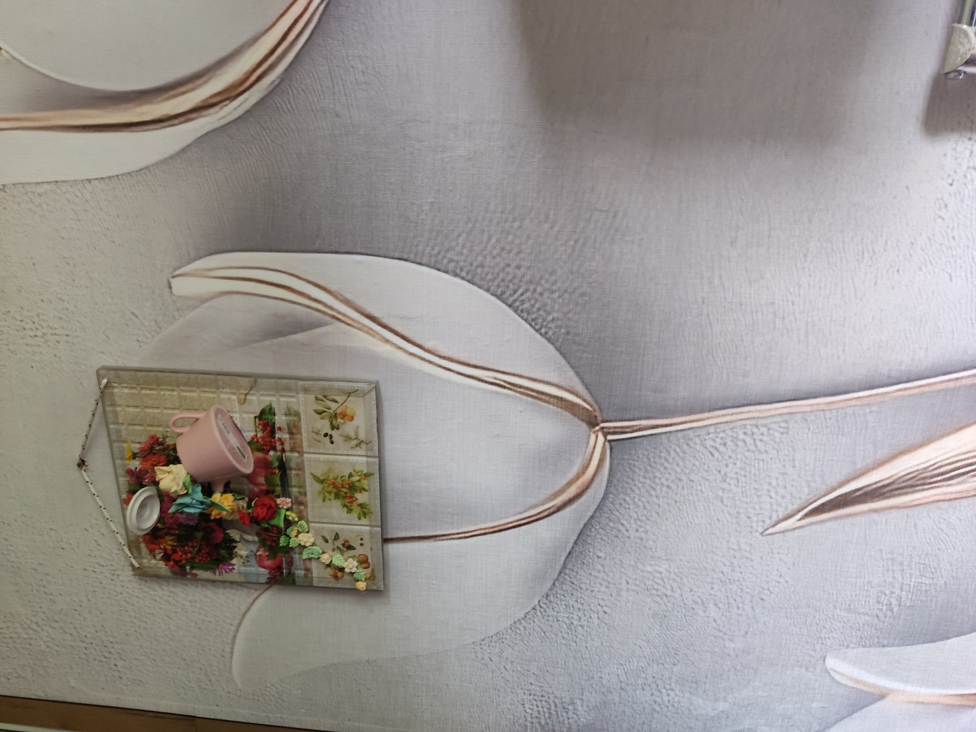 The image shows a close-up of a wall decorated with a relief of a large white tulip with copper accents. In the center of the tulip, a framed decorative panel is attached to the wall. This panel contains a miniature scene with a pink teacup tipped over a bouquet of colorful artificial flowers and berries. Below the bouquet are four small square tiles, each depicting a floral or fruit motif. The wall itself has a textured gray finish. No people are visible in the image. The location is not specified within the image or its crops.