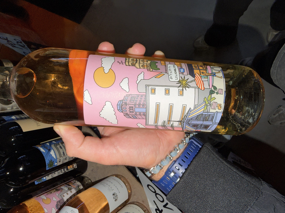 A person's right hand holds a transparent glass bottle containing an amber-colored liquid. The bottle features a detailed, stylized illustration on its label, set against a pastel pink background with a yellow sun and white clouds. The illustration depicts a street scene with various buildings, including a prominent multi-story structure and one with a clock tower, palm trees, and street-level elements such as a fruit stall (marked "1920"), potted plants, and a person walking under an umbrella. A street sign on the label clearly reads "לילינבלום" and "LILIENBLUM 46", indicating the specific location as Lilienblum Street in Tel Aviv, Israel.
The hand holding the bottle wears a dark long-sleeved garment, a blue digital watch on the wrist, and a light blue braided bracelet. In the background, numerous other wine bottles are visible on shelves, suggesting a retail environment such as a wine shop. One partially visible label on a pink-hued bottle reads "LA PERLE PINOT NOIR". A handwritten white sign, possibly indicating a price or promotion, shows "FOX 100" in the upper right background. The lower portion of the image reveals the person's legs and feet, clad in light-colored shoes, standing on a dark surface.