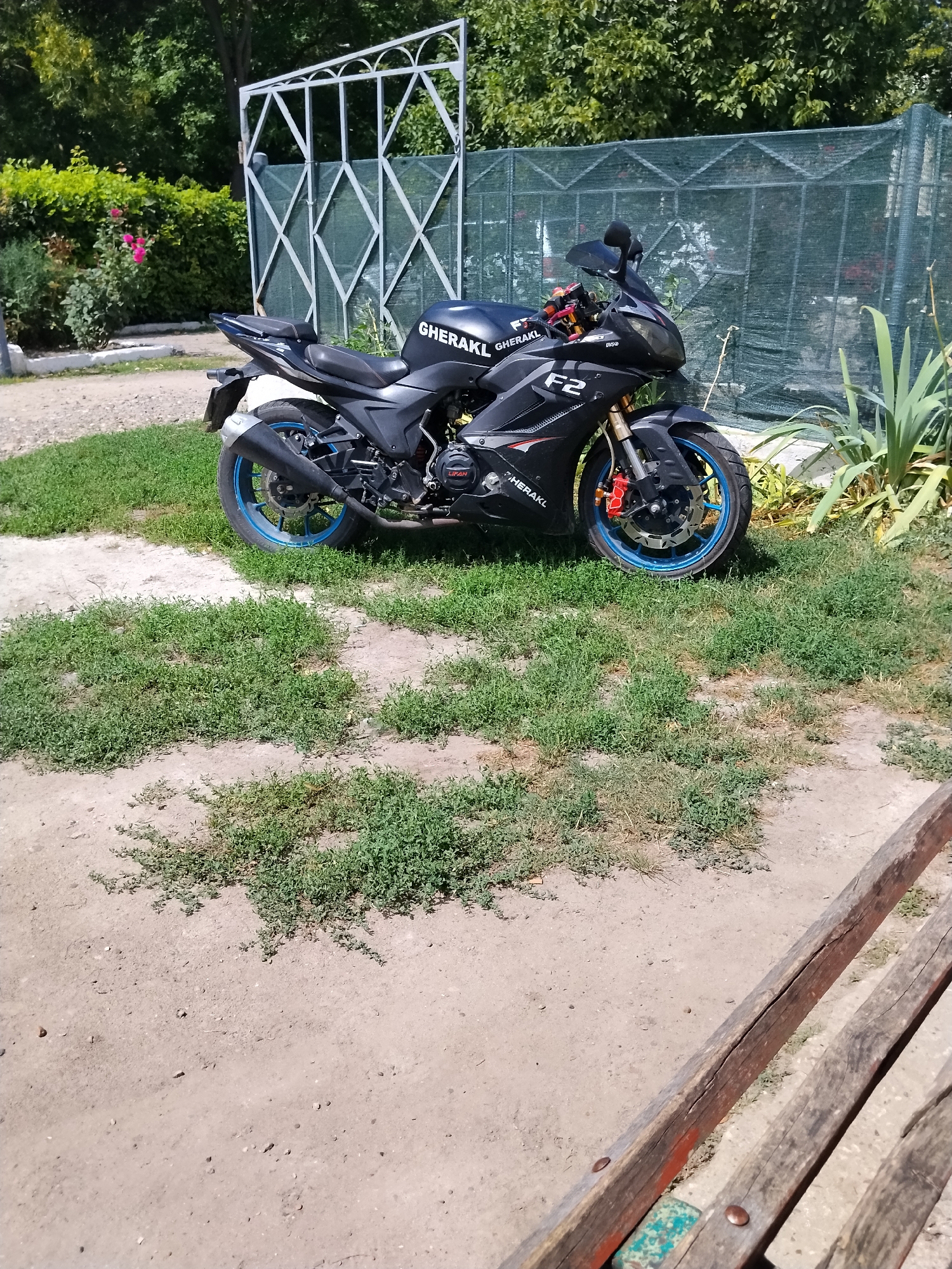 A black sport motorcycle is parked on a dirt and grass area in front of a green mesh fence and a white metal gate. The motorcycle has blue rims on its wheels and red brake calipers. The brand name "GHERAKL" is visible on the side of the motorcycle, along with the model designation "F2". In the background, there are green trees and bushes, with pink roses on a bush to the left. Part of a wooden bench is visible in the foreground, on the right side of the image. The scene appears to be outdoors in a rural or semi-rural setting.