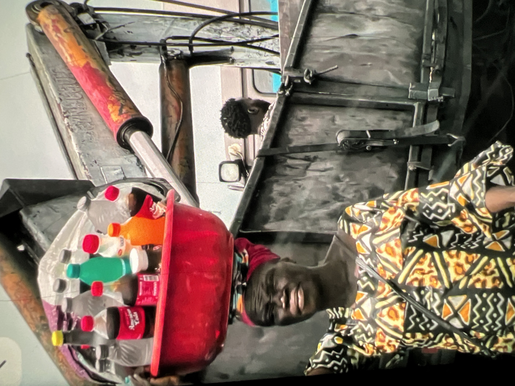 The image depicts a scene in Lagos, Nigeria, focusing on a woman balancing a red basin filled with various bottles of drinks and containers on her head. She wears a colorful, patterned dress and a red headscarf. Behind her, a man with dark, curly hair is visible from the side, looking away from the camera, seemingly inside a vehicle. The background includes industrial elements, such as a large metal truck or container with visible hydraulic machinery and a rusted, painted boom. The environment appears to be an urban setting, possibly a street or a loading area, characterized by industrial equipment and a somewhat gritty atmosphere. The lighting suggests an outdoor environment during the day.