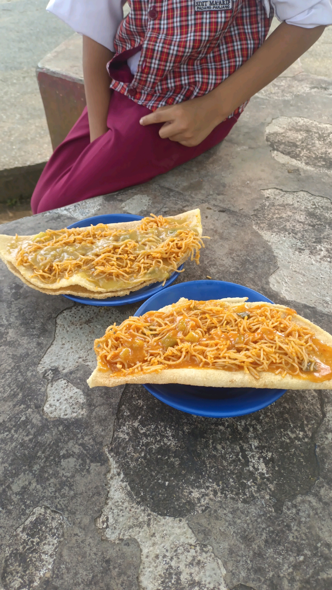 Two blue plates hold crispy, folded food items topped with a savory noodle mixture. The plates rest on a textured, grey outdoor surface. In the background, a person wearing a red and white plaid vest over a white shirt and maroon pants is partially visible, sitting on a concrete ledge. A visible label on the vest reads "SDIT MA'ARIF PADANG PANJANG" indicating the location as a school in Padang Panjang, Indonesia. The overall scene suggests a casual outdoor setting, possibly a schoolyard or common area.
