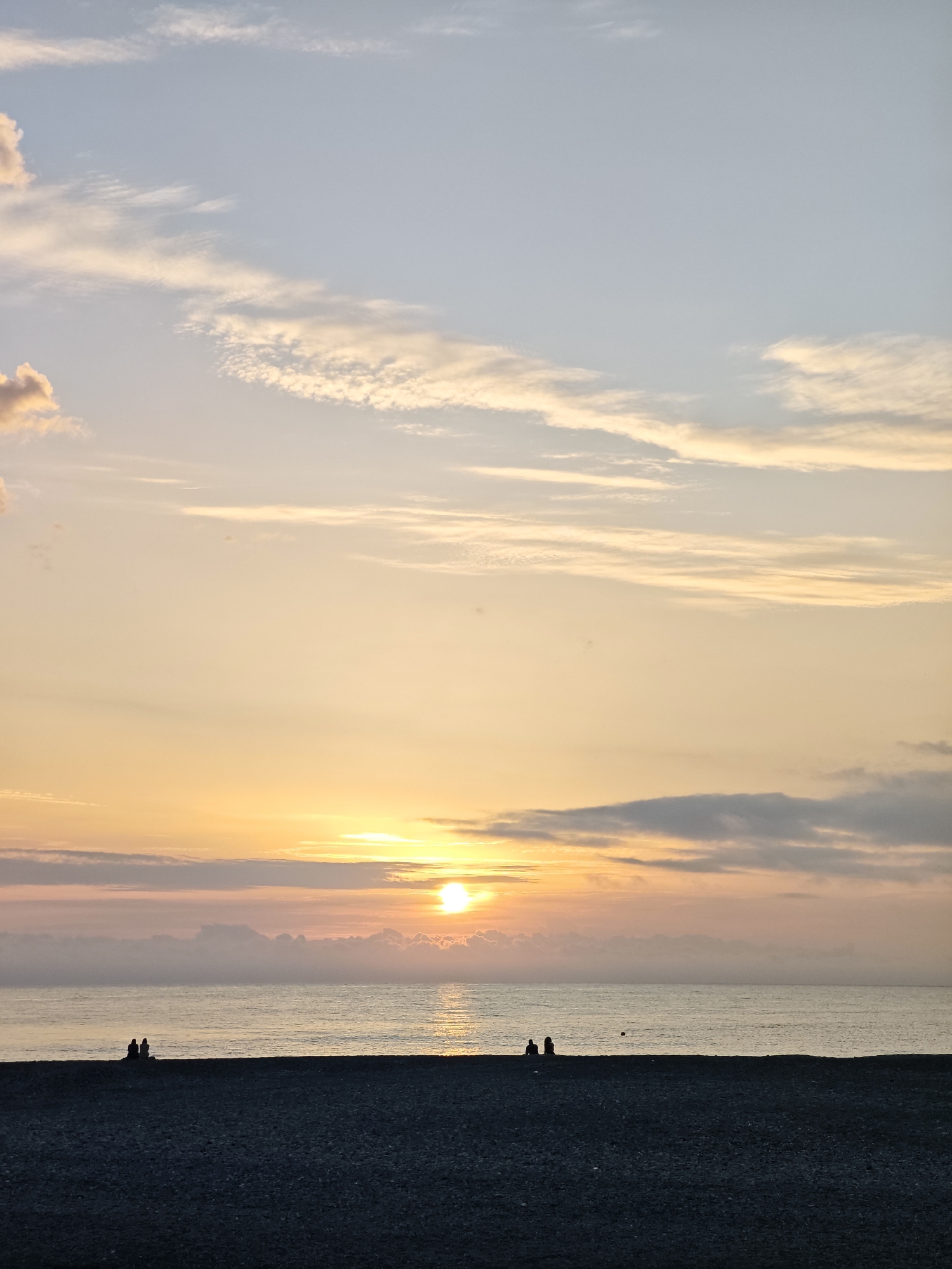 The image shows a seaside scene in Sochi, Russia at sunset. Two pairs of individuals are visible in silhouette on a pebble beach, with the sea stretching out to the horizon. The sun, partially obscured by clouds, casts a golden glow over the water and the sky, which is filled with pale clouds.