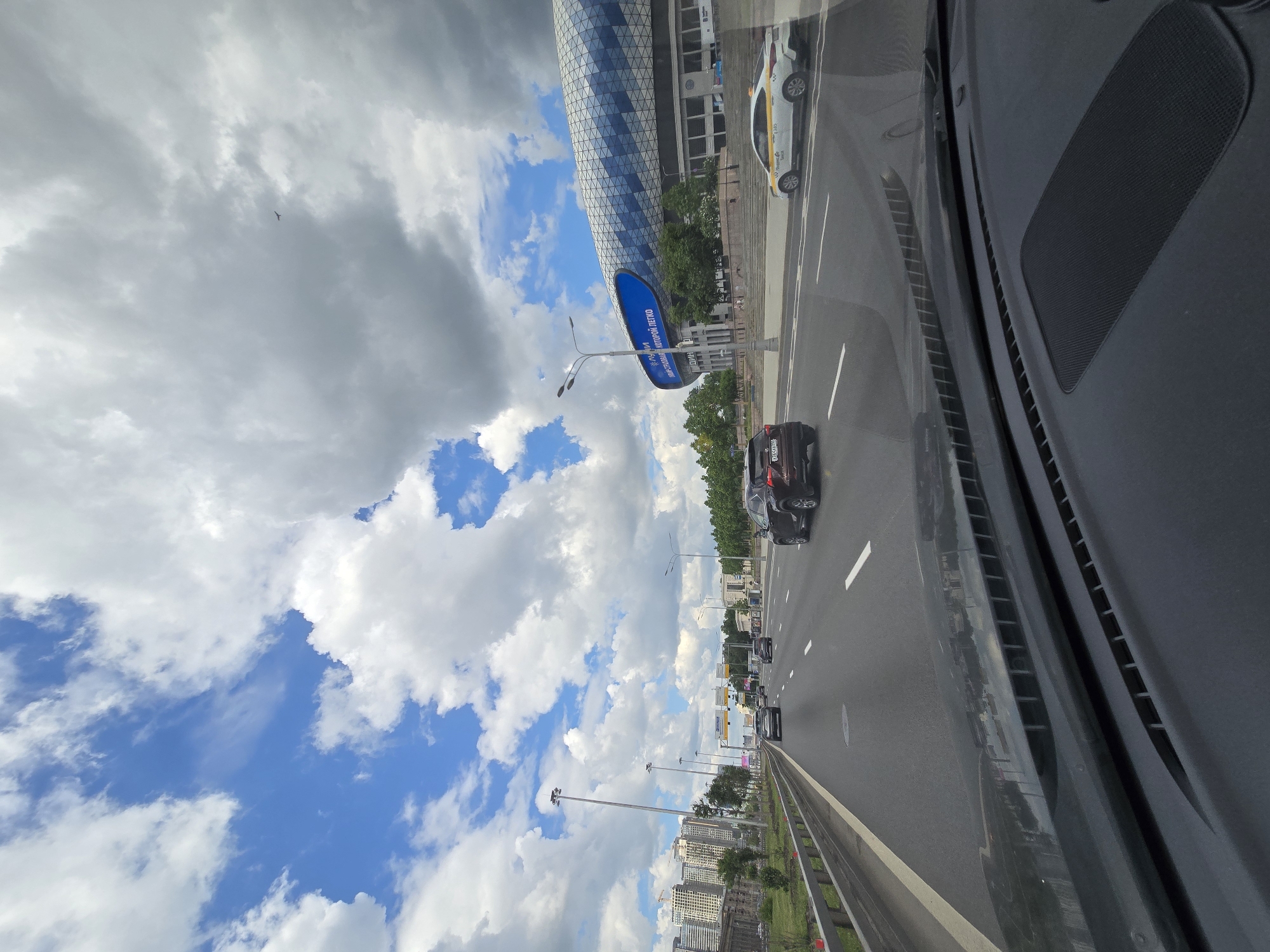 The image shows a view from inside a car, looking out onto a highway in Moscow, Russia. On the right, the vehicle's dashboard is visible. A multi-lane highway stretches out in front, with multiple cars traveling in both directions. A distinctive, cylindrical building with a geometric pattern is visible on the left side of the road, close to the roadway. There is also a large blue sign and various light posts. Beyond the road, there is green foliage including trees and grass. Further away, buildings are visible, including apartment buildings. The sky is filled with bright blue patches interspersed with fluffy white clouds.