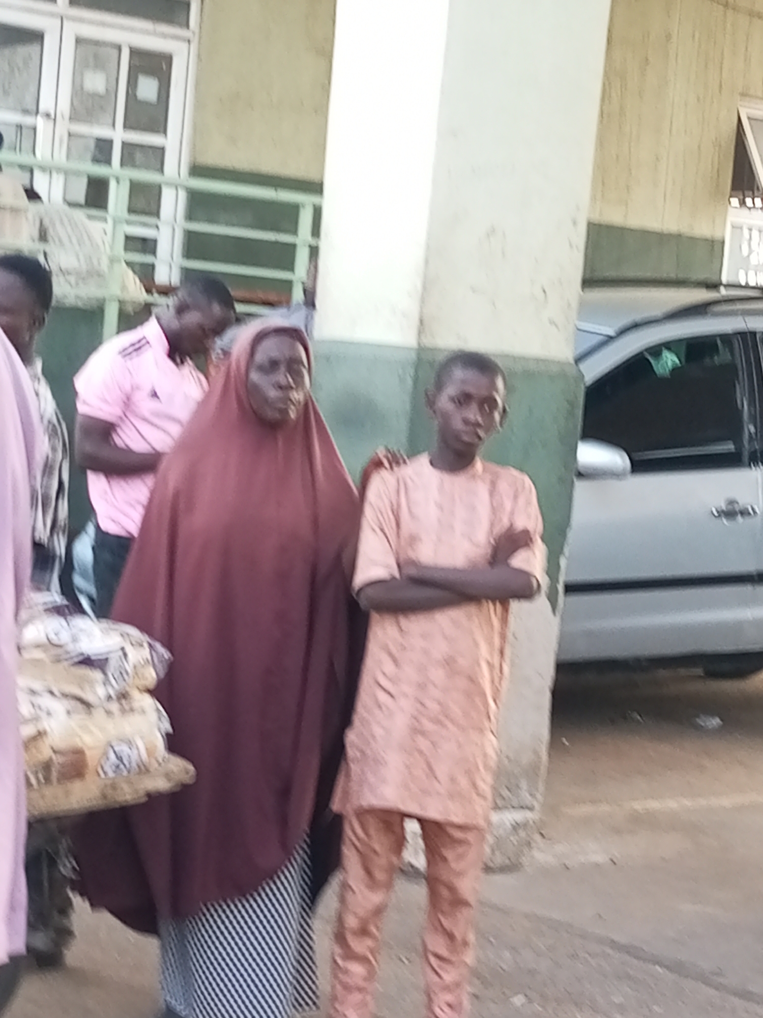 In Gombe, Nigeria, a group of people stand outside a building. In the foreground, a woman wearing a dark red hijab and a striped skirt stands next to a young boy in a light orange patterned tunic and pants. The boy has his arms crossed. To their left, a man in a pink shirt is partially visible. Behind them, a light-colored car is parked. The background shows a building with windows and a green railing.