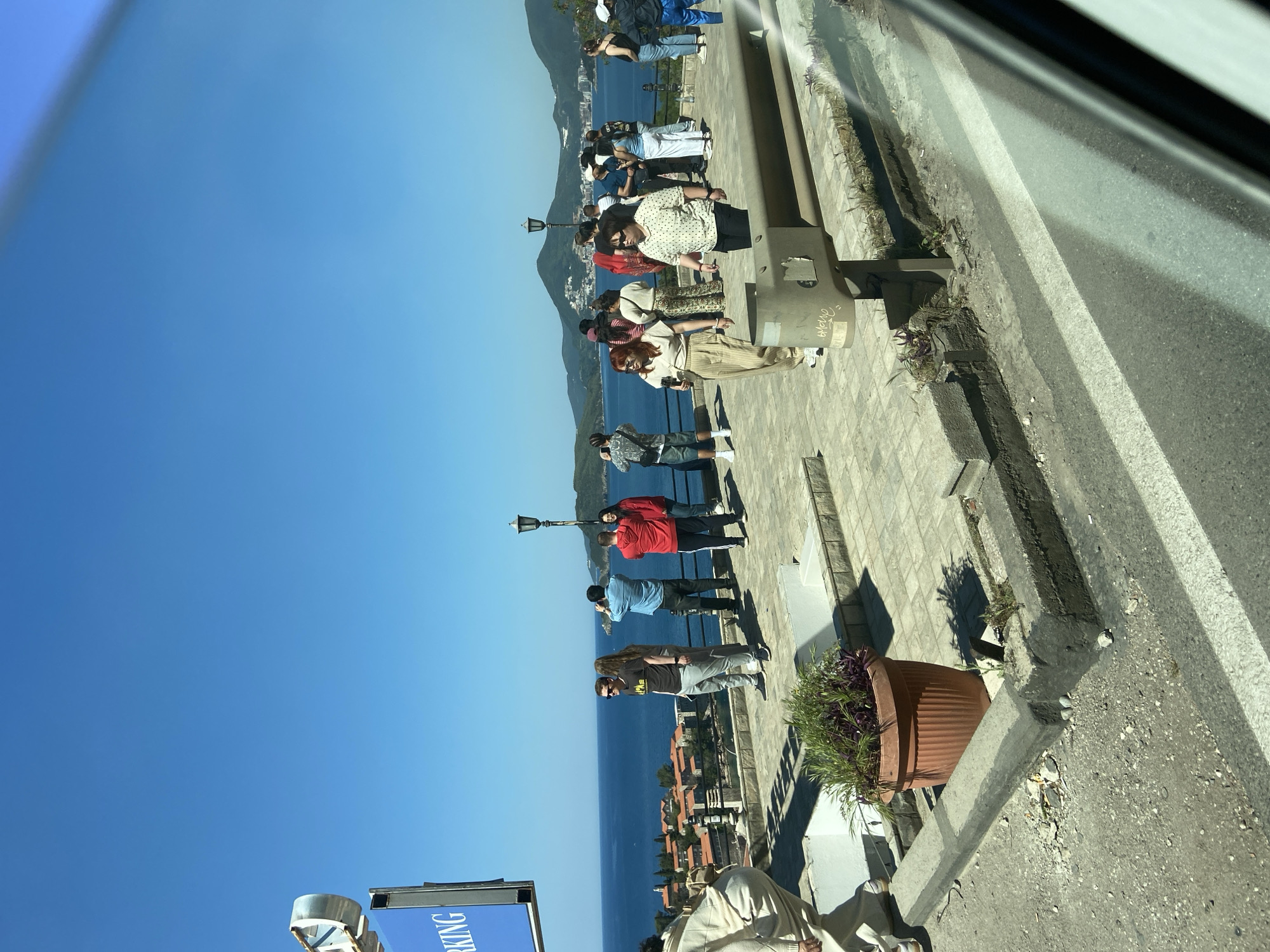 The image, taken from a slightly elevated position (likely from a vehicle), shows a view of the city of Sveti Stefan in Montenegro. The sea dominates the left side of the frame, with a clear blue sky above. Along the coast, a group of people, approximately ten, are standing on a paved walkway, many facing towards the sea and seemingly observing the scenery. Some people are wearing face masks. A few street lamps are visible along the walkway. In the foreground, partially cut off at the bottom, is a plant pot. To the right, the edge of a road is visible, with double white lines.