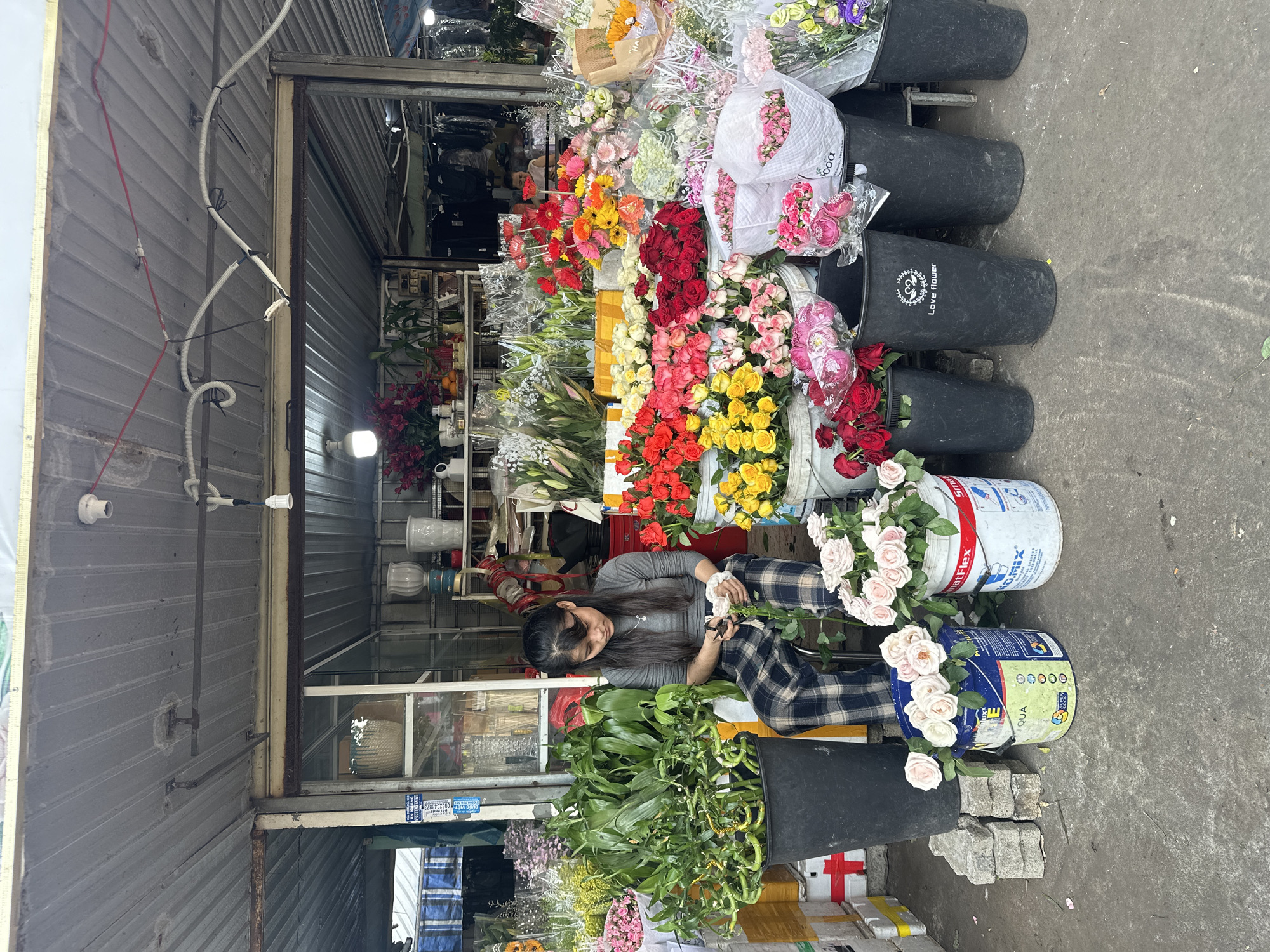 A woman is seen sitting in a flower stall in Đà Nẵng, Vietnam. She is wearing a gray long-sleeve shirt and checkered pants. She is arranging roses, cutting stems, and creating bouquets. The stall is filled with various flowers arranged in black buckets and on shelves. The environment is cluttered, but colorful due to the abundance of flowers. The storefront is made of corrugated metal with a glass display case inside.