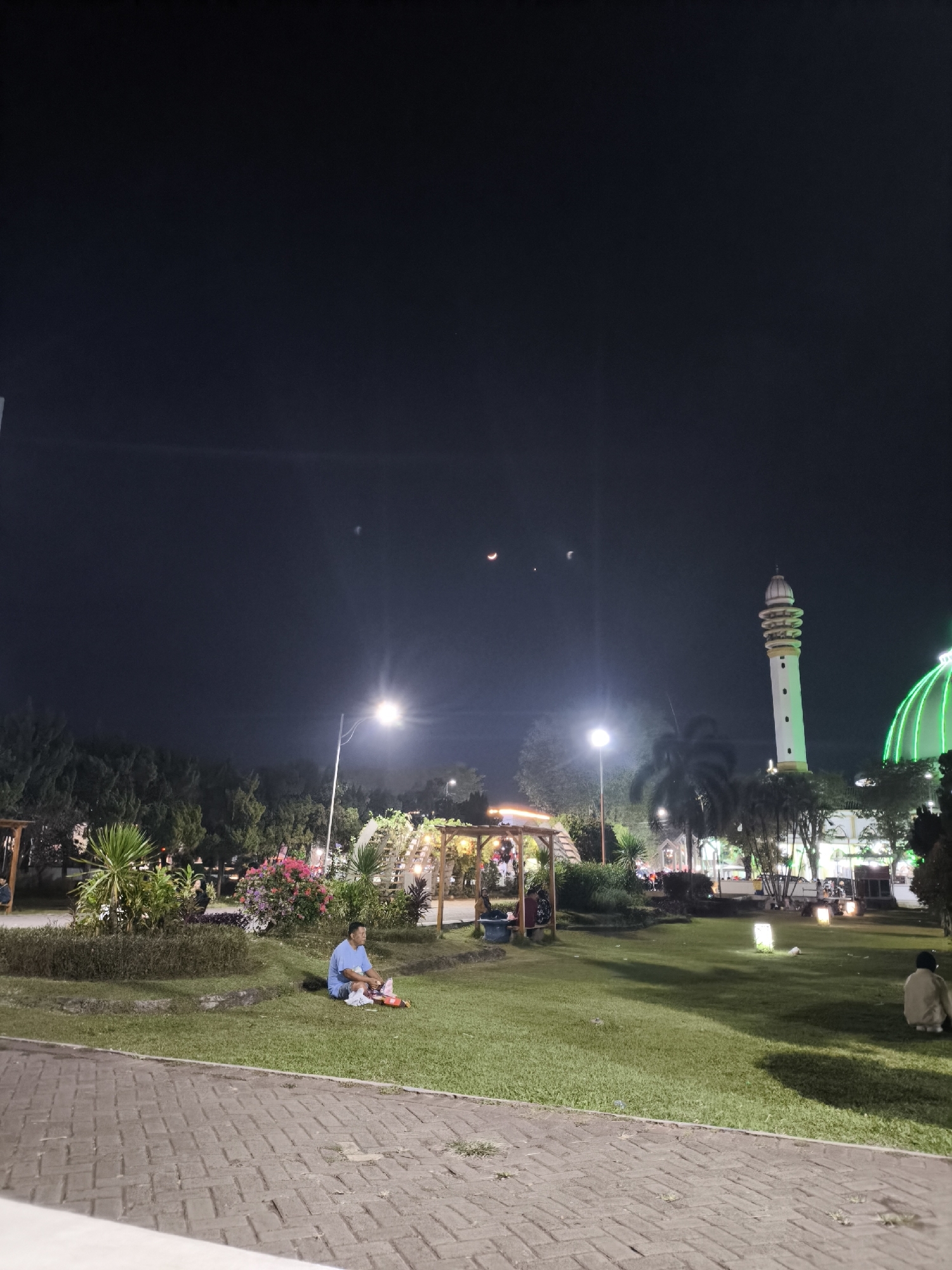 The image captures a night scene in what appears to be Alun-Alun Lumajang, a town square in Lumajang, Indonesia. The foreground is dominated by a paved walkway made of reddish-brown bricks. Beyond the walkway, a well-maintained lawn stretches across the scene. In the mid-ground, a man in a blue shirt and shorts is seated on the grass, facing away from the camera. He is looking at a red mat laid out before him, on which there are some items, possibly food. To his right, a garden bed features a variety of plants, including green shrubs and vibrant pink flowers. Further back, a wooden gazebo structure is visible, illuminated by warm lights.
To the right of the gazebo, a minaret of a mosque rises tall and slender, with a white and yellow exterior, illuminated by green lights on the mosque's dome. The mosque dome is a prominent feature, glowing with green light. In the dark night sky above, three celestial bodies are visible: two crescent moons of varying sizes and a small, circular celestial object, possibly a planet or a star, positioned between them. Several streetlights are also scattered throughout the scene, casting pools of light. In the far right background, another person is seen sitting on the grass, facing away from the camera. The overall atmosphere is serene and communal, suggesting a public park or gathering space at night.