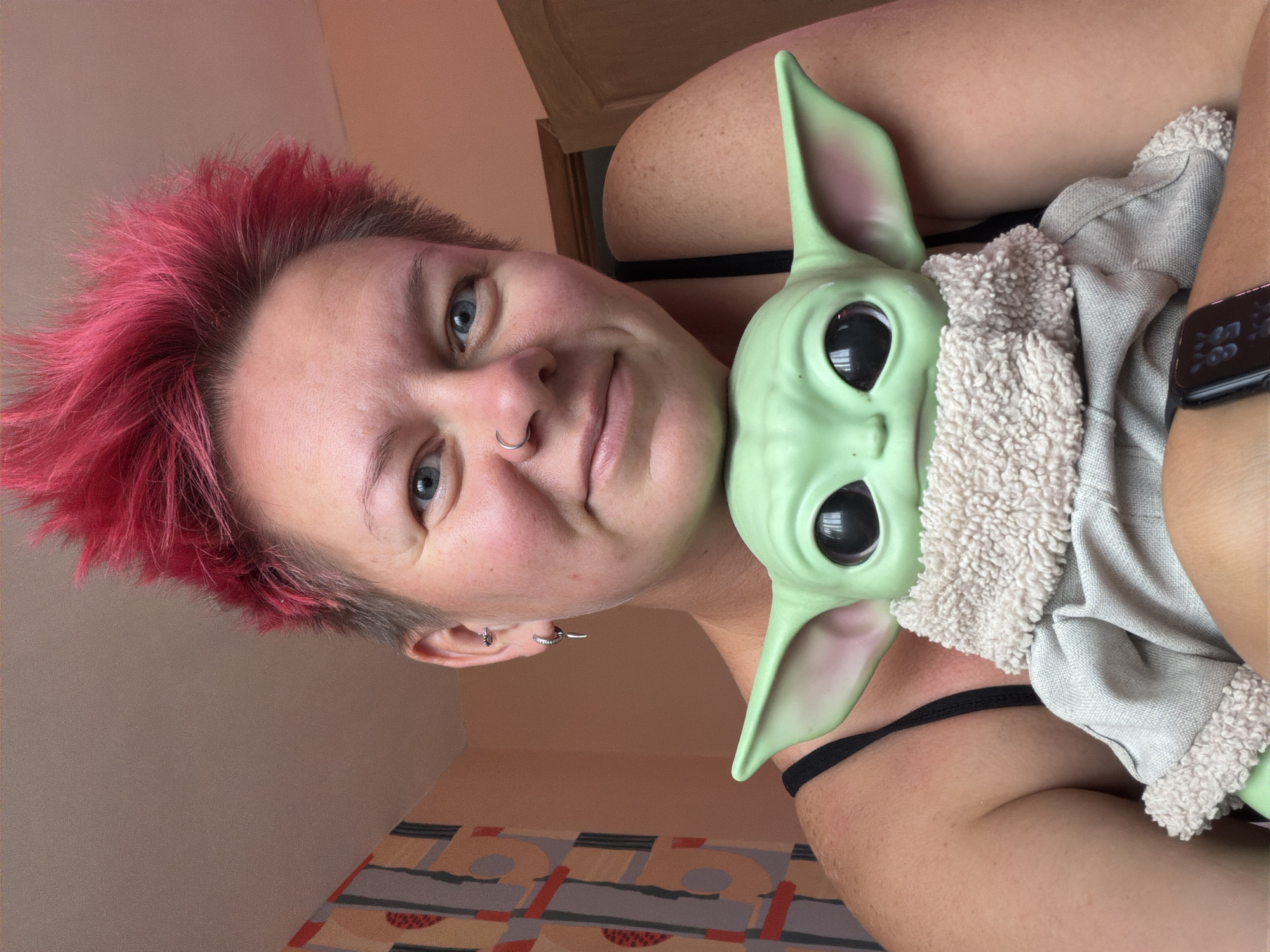 A close-up, selfie-style photograph captures a person with short, spiky, bright pink hair and blue eyes, holding a plush toy of Grogu (Baby Yoda). The person is wearing a black tank top and has silver hoop earrings and a silver nose ring. The Grogu toy is wearing its signature robe. The background appears to be an indoor setting with a peach-colored wall and a portion of patterned wallpaper visible on the left. The image is taken from a slightly high angle, looking down at the person and the toy. There is no indication of the city of Chaikovsky, Russia, in the image.