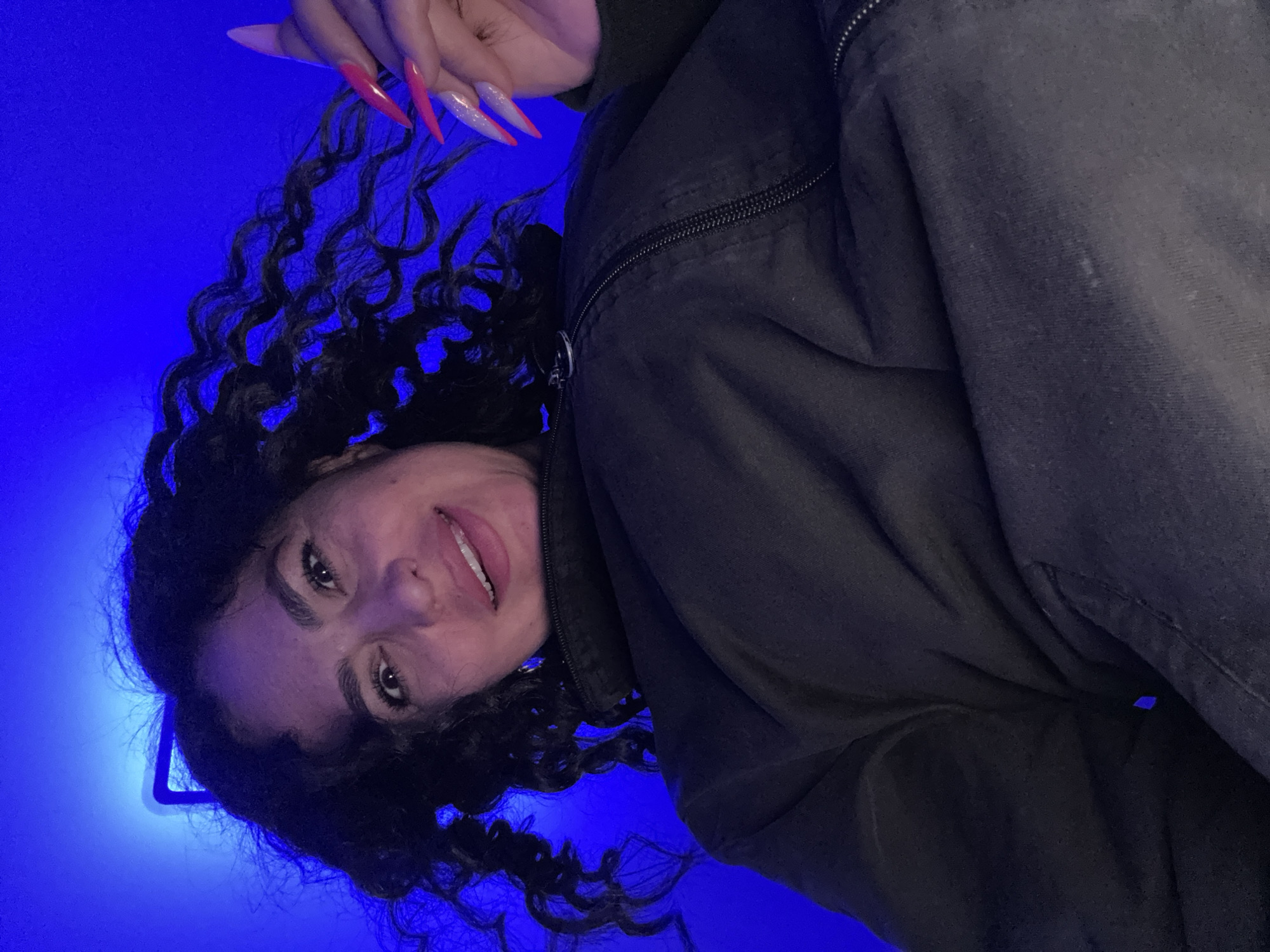 A woman with curly dark hair, light skin, and long pink and white fingernails is wearing a dark jacket. The background is a bright blue color. The image seems to be taken from a low angle and is slightly rotated. There are no indications of the location being Kauchuk Stantsiya, Russia.