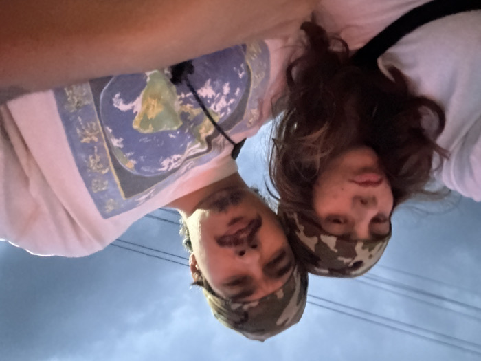 A close-up, low-angle photograph features two individuals outdoors in Ban Mai Saha Sampan, Thailand, against a dull blue-grey overcast sky with faint power lines visible. The individual on the left has long, wavy dark brown hair, wears a camouflage pattern headband, and a white t-shirt, with a black strap visible over their shoulder. Their lips are slightly pursed. The individual on the right, partially visible, has a mustache and short dark hair, also wearing a camouflage pattern headband. They wear a white t-shirt with a prominent blue graphic of the Earth, accompanied by East Asian script. A black cord is visible around their neck. Both individuals are positioned closely together, looking towards the camera.