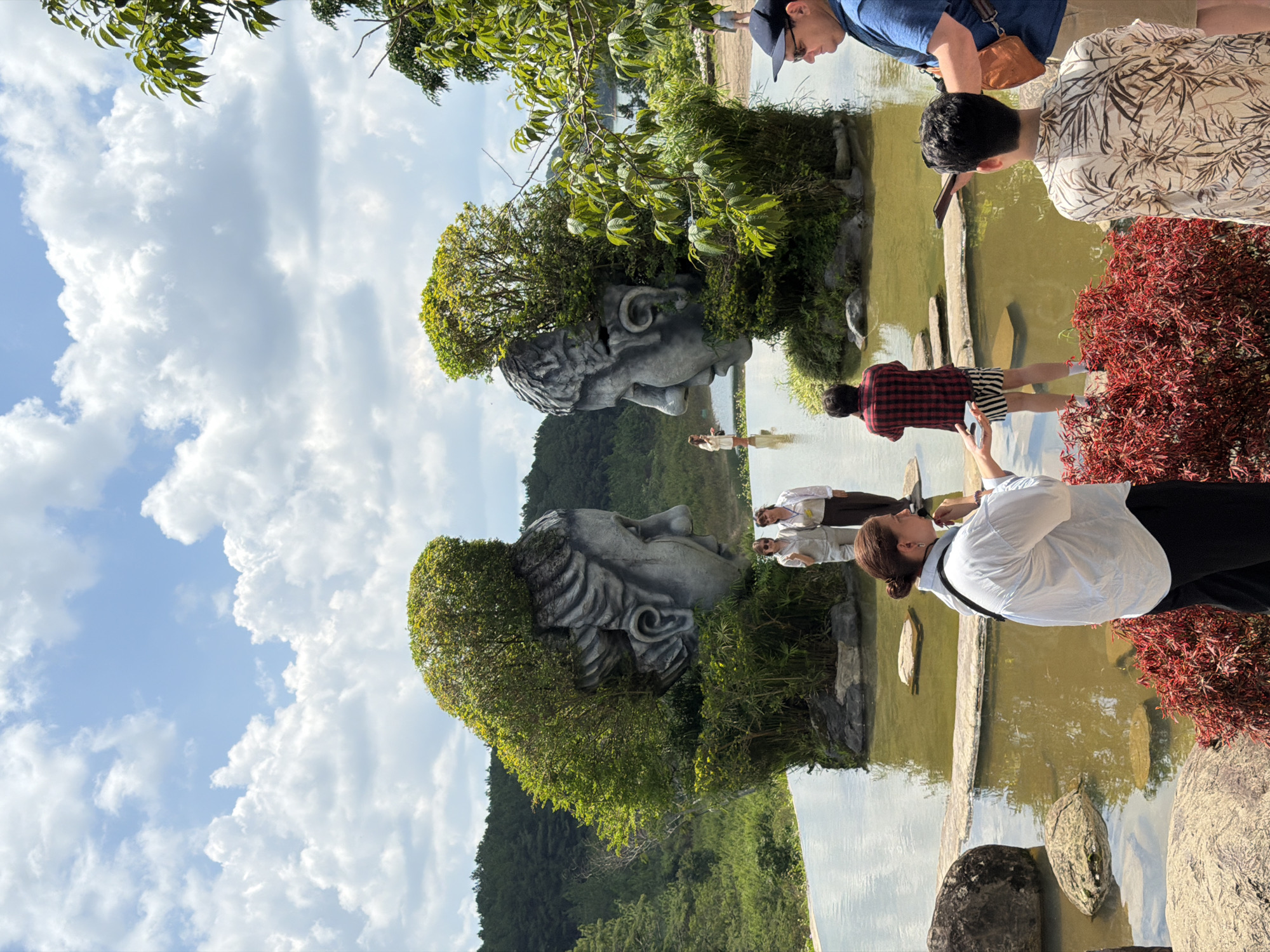 This is an outdoor photograph taken in the city of Da Lat, Vietnam, likely at the Da Lat Wonderland theme park or a similar attraction featuring artistic sculptures. The central focus is on two large, stone sculptures of human heads, positioned facing each other across a shallow, reflective body of water. These heads are artistically integrated with greenery, with trees and bushes growing out of their tops and sides, creating a surreal, naturalistic effect.
In the foreground, several people are present, appearing to be tourists. On the left, a woman with her back to the camera is wearing a white shirt and black pants, holding a phone or camera and possibly gesturing. Next to her, slightly further back, a person in a red and black checkered shirt and striped shorts is looking towards the sculptures. To the right of the frame, a man wearing a blue shirt and a dark baseball cap is visible, and to his right, another person in a light-colored, patterned shirt is also facing away from the viewer.
The water in front of the sculptures has stepping stones or decorative rock formations placed within it. In the background, a lush green landscape of hills and trees stretches to the horizon, under a bright sky with scattered white clouds. The overall impression is a scenic, somewhat whimsical tourist destination.