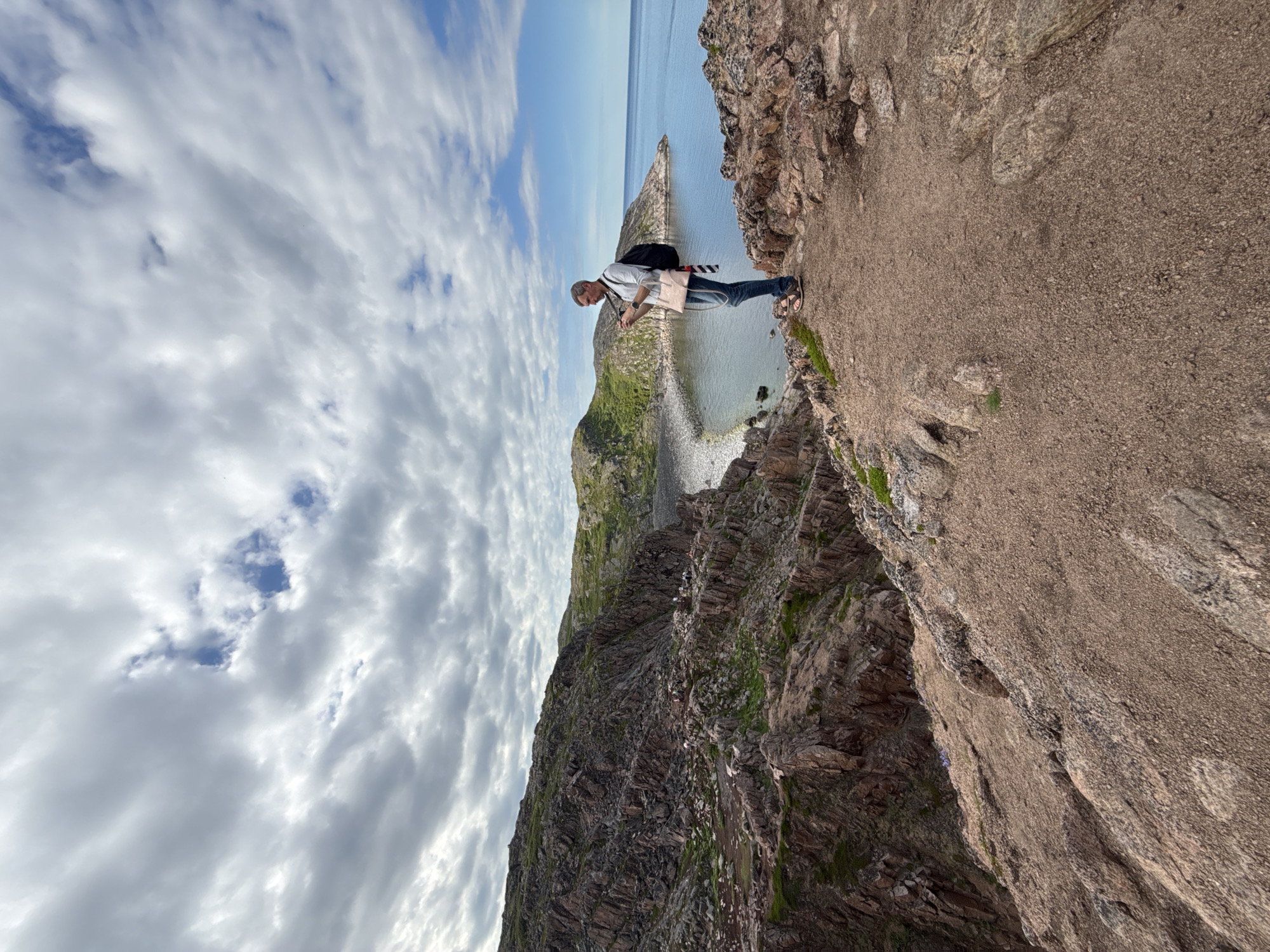 A full-shot, low-angle, outdoor, natural light photograph captures a man standing on a rocky cliff edge overlooking the Barents Sea. The man, with short, graying hair, is wearing a light-colored long-sleeved shirt, blue jeans, and sandals. He has a dark backpack slung over his left shoulder and a light-pink cross-body bag over his right. He is holding a camera up to his face, seemingly taking a picture of the scenery. The cliff is made of rough, reddish-brown rocks with patches of green moss and grass. Below the cliff, a rocky beach leads to the calm, blue water. In the distance, rolling green hills rise from the coastline under a cloudy sky with patches of blue. Several other people are visible on a lower ledge of the cliff, also appearing to be observing the view. The location is the coast of the Arctic Ocean in Teriberka, Russia.