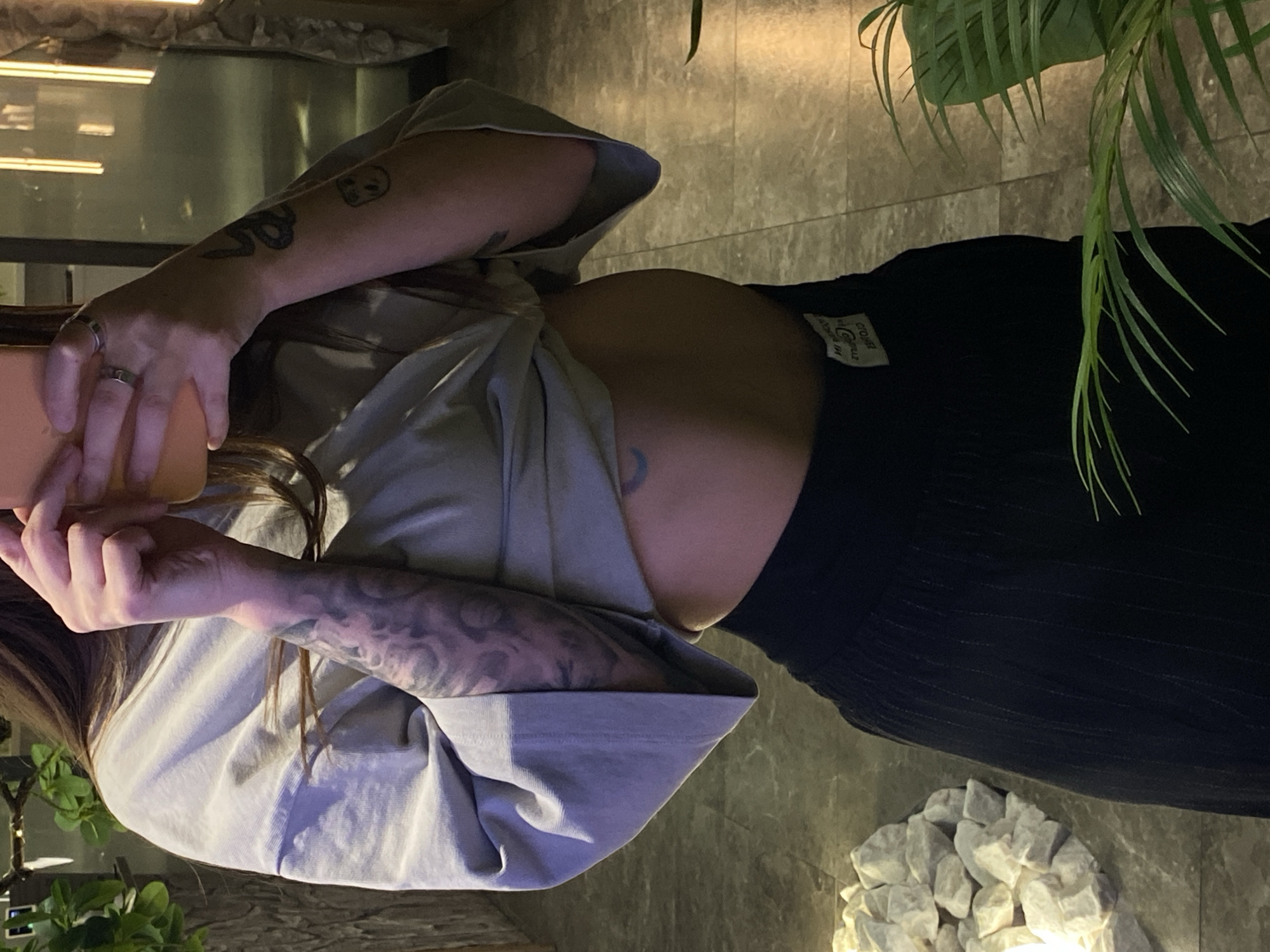 A person stands in an indoor setting, holding a mobile device, possibly taking a photograph or viewing content on the screen. The individual is wearing a light grey, oversized t-shirt and black pants. Tattoos are visible on their arms, including a snake and a skull. A small, crescent moon tattoo is also visible on their abdomen. There are plants in the background, and a pile of white stones is situated on the floor. The location is unknown, but the prompt indicates it may be in the city of Votkinsk, Russia.