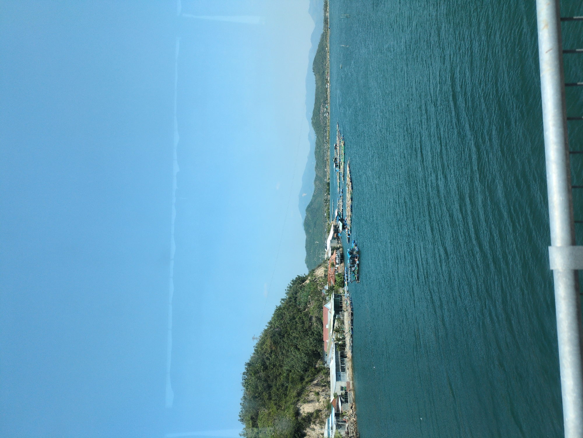 The image captures a view in Hòa Mỹ, Vietnam, showing a coastline adjacent to a body of water. The scene is divided vertically into a clear blue sky on the left and dark turquoise sea on the right. A narrow strip of land separates the sky and the sea. The land features a mix of dense green vegetation and residential structures, some with visible rooftops. Several boats are docked along the coast. The sky has minimal cloud cover, and the sea appears to have slight surface ripples. A portion of what seems to be a metal structure or support pole is present on the right edge of the image.