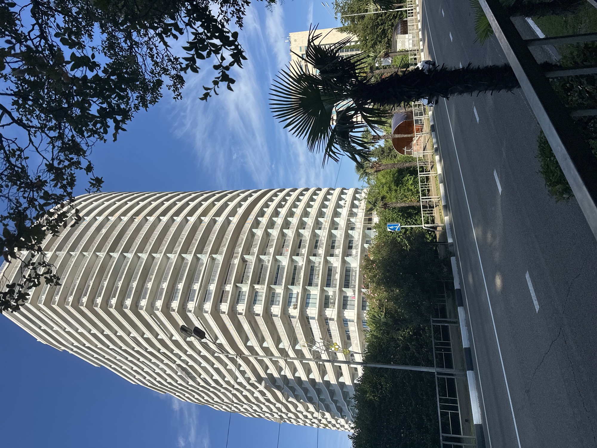 The image shows a view of a curving, multi-story apartment building with many balconies, likely located in Sochi, Russia. The building is white and appears to be modern. In the foreground, there is a road with a white line marking the lanes and a sidewalk. A tall palm tree stands in front of the building, and other trees and greenery are visible. The sky is blue with some wispy clouds. In the background, another lighter-colored building and some more trees can be seen. A streetlamp is visible near the apartment building. A blue pedestrian crossing sign is present on the left side of the road. A car is driving on the road. The overall scene suggests a sunny day in a coastal or resort area.