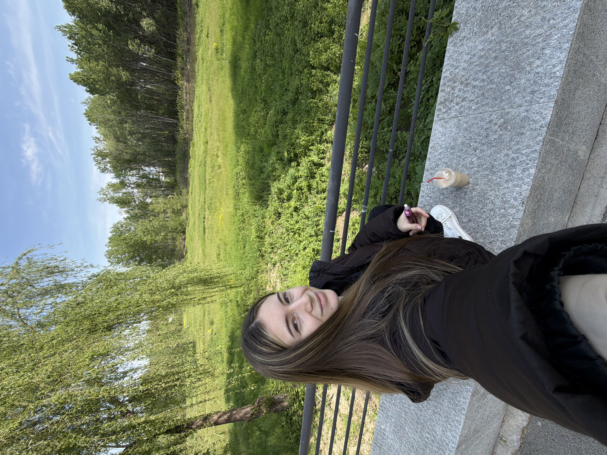 A young woman is captured in a vibrant outdoor setting, appearing relaxed as she poses for the camera. She is visible from the chest up, with her long, dark brown hair featuring lighter highlights cascading over her shoulders. She looks directly forward with a gentle, pleasant smile. She wears a black jacket, and parts of her white sneakers are visible towards the bottom of the frame.
She is situated next to a sturdy gray, granite-like stone ledge or wall, which also features a black metal railing running along its edge. On this stone surface, a clear plastic cup with a red straw, containing a light brown beverage (likely an iced coffee), rests. Her right hand, partially visible, holds a small pink and white pen or marker.
The background unfolds into a lush natural landscape under a bright, clear blue sky with a few wispy white clouds. A wide expanse of vivid green grass and foliage dominates the mid-ground, stretching towards a dense line of tall, leafy trees in the distance. Closer to the foreground, a prominent tree with long, drooping branches and bright green leaves, resembling a weeping willow, adds depth to the scene. The lighting suggests a sunny day, likely late morning or afternoon.
The overall atmosphere is serene and natural, suggesting a leisurely moment in a park or by a riverside, such as those found in Plovdiv, Bulgaria. No visible text is present in the image.