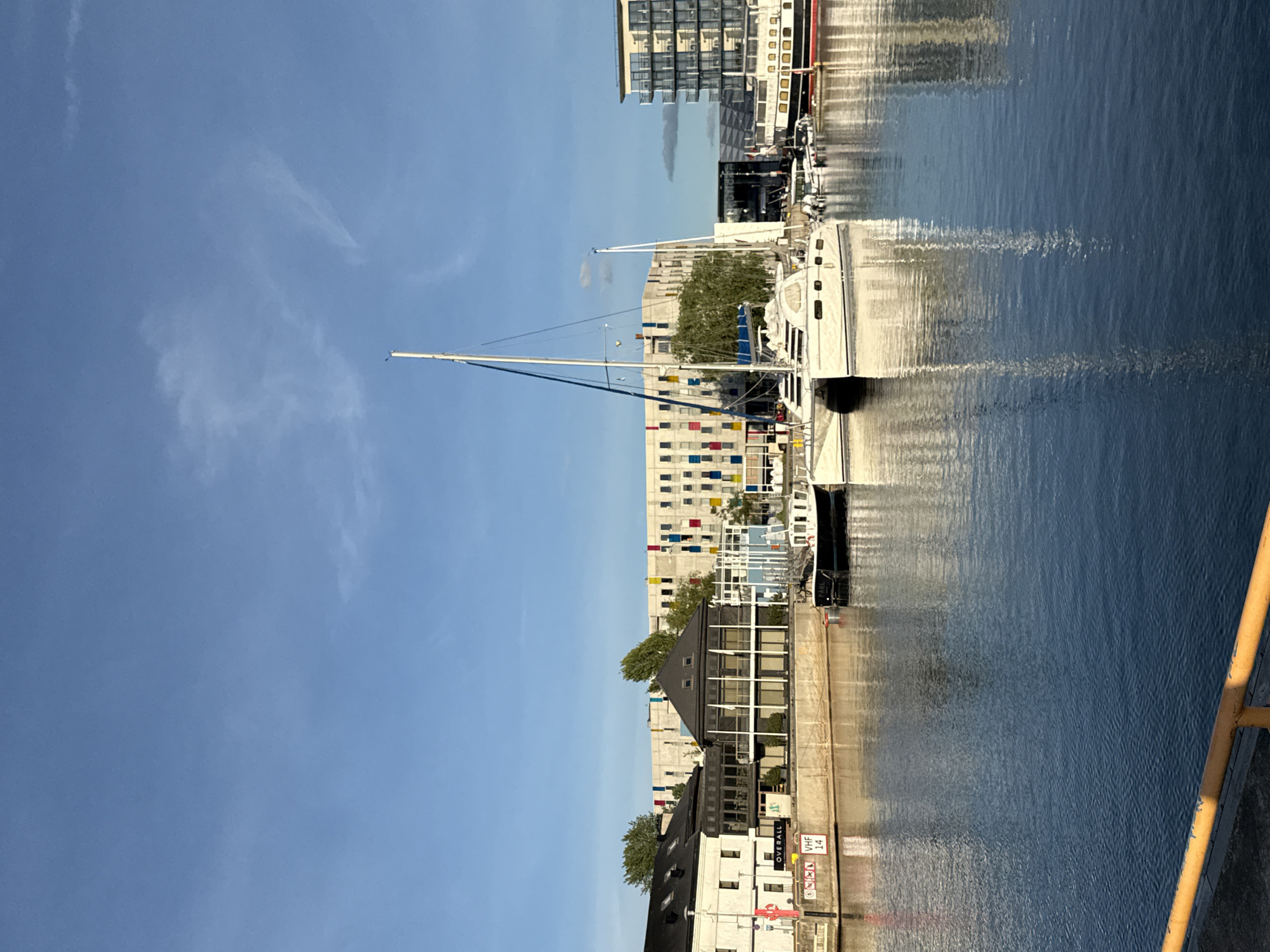 A waterscape view is captured in Tallinn, Estonia, featuring a calm body of water reflecting the surrounding architecture and boats under a clear blue sky with a few wispy clouds. In the foreground, a yellow railing is visible along the edge of what appears to be a dock or promenade. Several boats are moored in the water, including a white catamaran with its sails partially furled and a smaller white boat docked beside it. Further in the background, there are various buildings. One building is a modern structure with a distinctive facade featuring rows of colorful rectangular panels in red, yellow, blue, and green. Adjacent to this is a building with dark pitched roofs and large windows. Another building to the far right is a multi-story structure with a glass facade and balconies. The scene suggests a harbor or marina area within the city. There are no people clearly visible in the image. The lighting indicates it is likely daytime, possibly morning or late afternoon, given the angle of the sun and the soft shadows.
