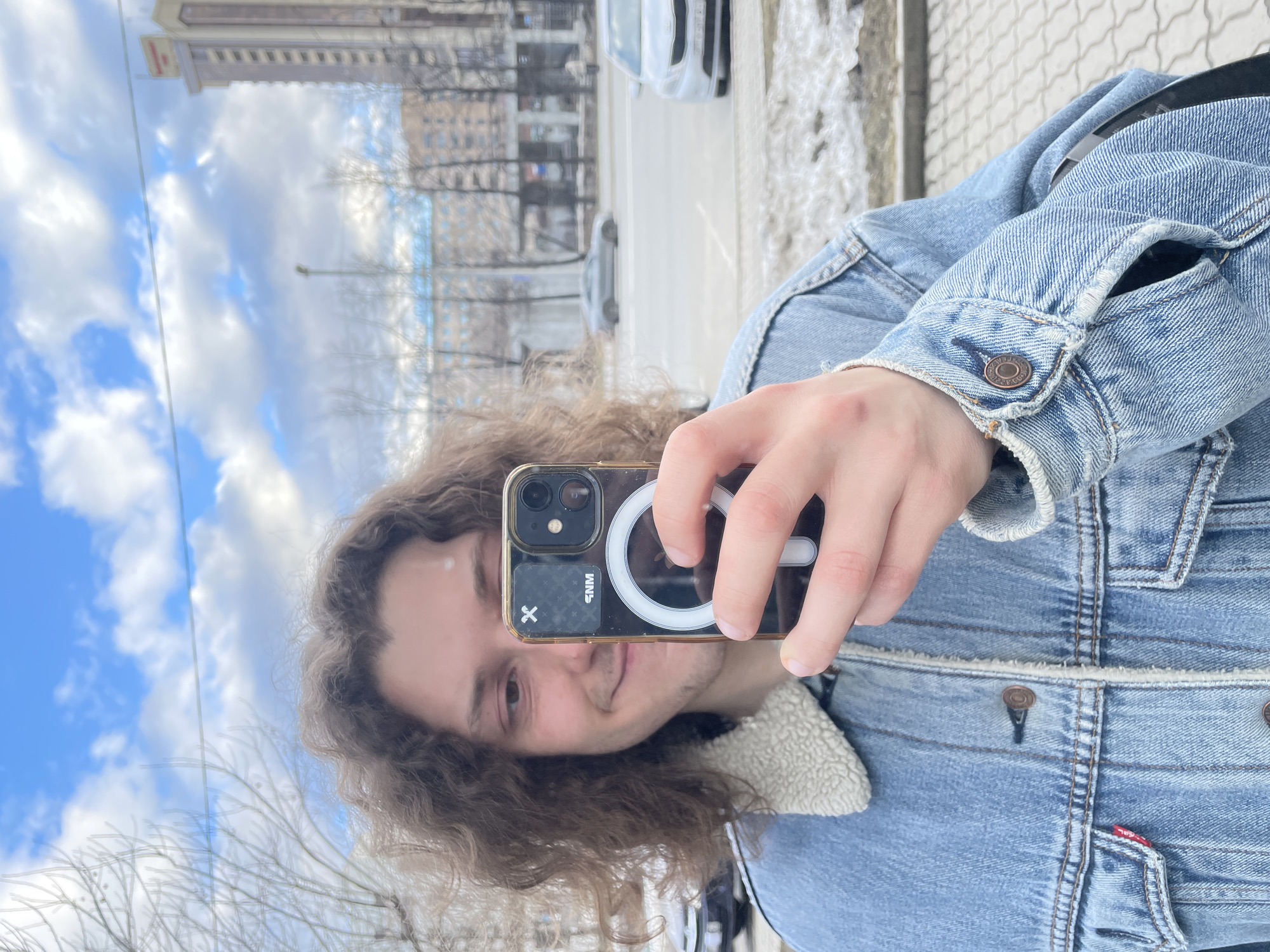 A person with long, curly hair is taking a selfie outdoors. They are holding an iPhone with a clear case and a MagSafe charger attached to the back. They are wearing a blue denim jacket with a shearling collar. The background includes a city street with buildings, trees, and snow on the ground. A tall building is visible in the background of the photo, which suggests this picture was taken in Yekaterinburg, Russia, with consideration to the skyline structure. The sky is partly cloudy with blue patches. A car is visible parked on the street in the background. The person's face is partially obscured by the phone, but they appear to be smiling slightly. The image is taken in portrait mode and the camera is held at eye level. The paved sidewalk texture is visible near the road.