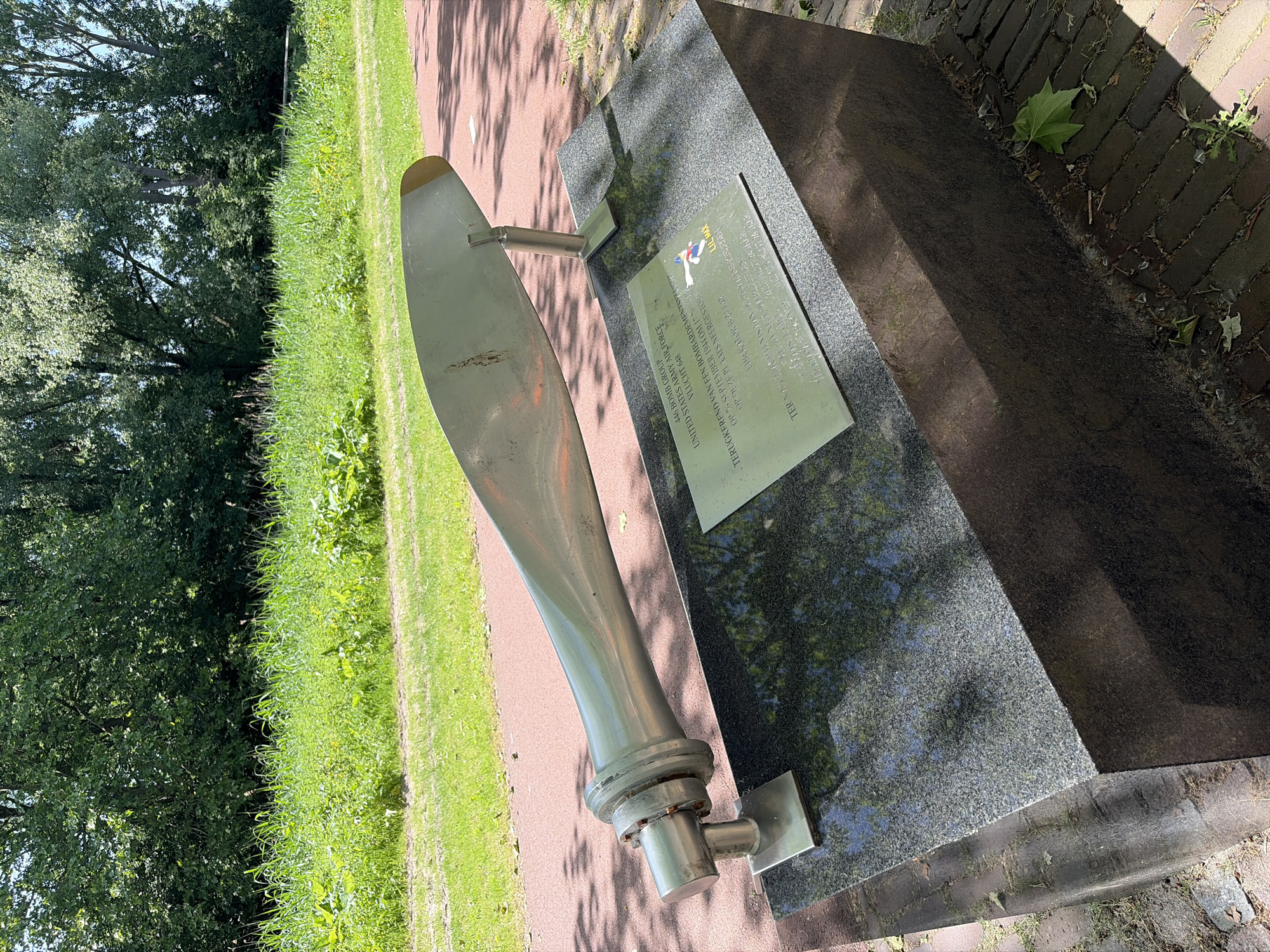 The image shows a memorial dedicated to the crew of the Liberator B-24 "Lil Max" that crashed in Rijswijk, Netherlands. The memorial consists of a polished aircraft propeller mounted on a stone base with a plaque. The plaque commemorates the crew who died during a bombing mission on September 26, 1944. The memorial is located in a park-like setting with grass and trees in the background.