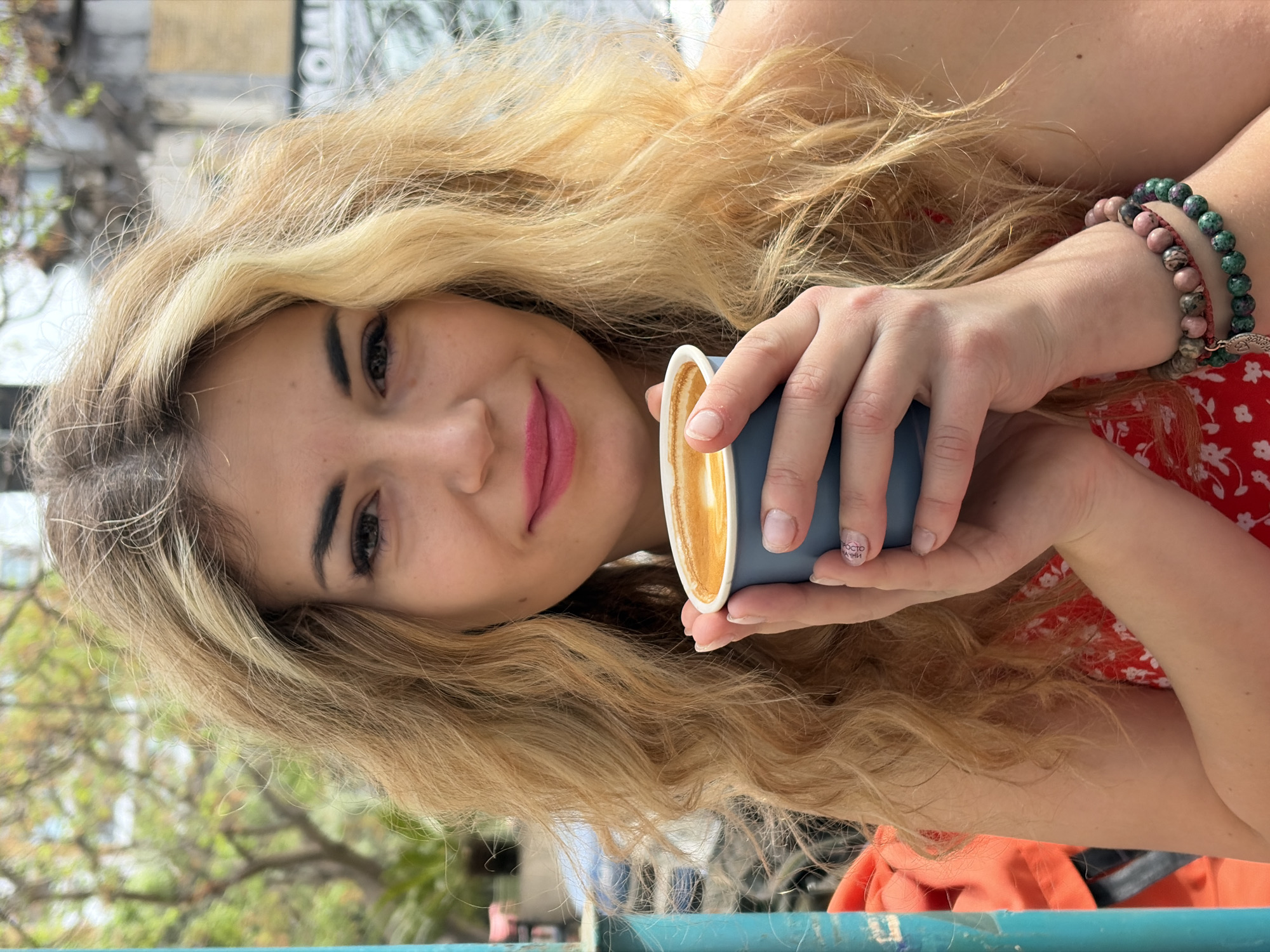 A young woman with long, wavy, blonde hair holds a small blue cup of coffee. She is wearing a red floral dress and has two beaded bracelets on her right wrist. Her lips are pink, and she appears to be smiling slightly at the camera. The background is blurred, suggesting an outdoor setting possibly in Budva, Montenegro, as evidenced by the possible architectural elements and greenery visible. She might be sitting at a table, as indicated by a blue, metallic structure visible in the foreground.