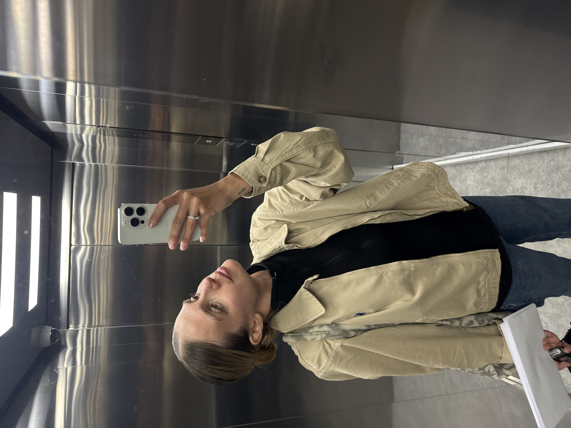 A woman is taking a selfie in an elevator. She is holding a white smartphone in her right hand and looking at the screen. The woman has light brown hair pulled back in a bun and is wearing a beige jacket over a black top and blue jeans. She is holding a stack of white papers in her left hand. The elevator walls are silver and shiny, reflecting the light. A security camera is visible in the upper left corner of the elevator. The location is Unknown, Unknown.