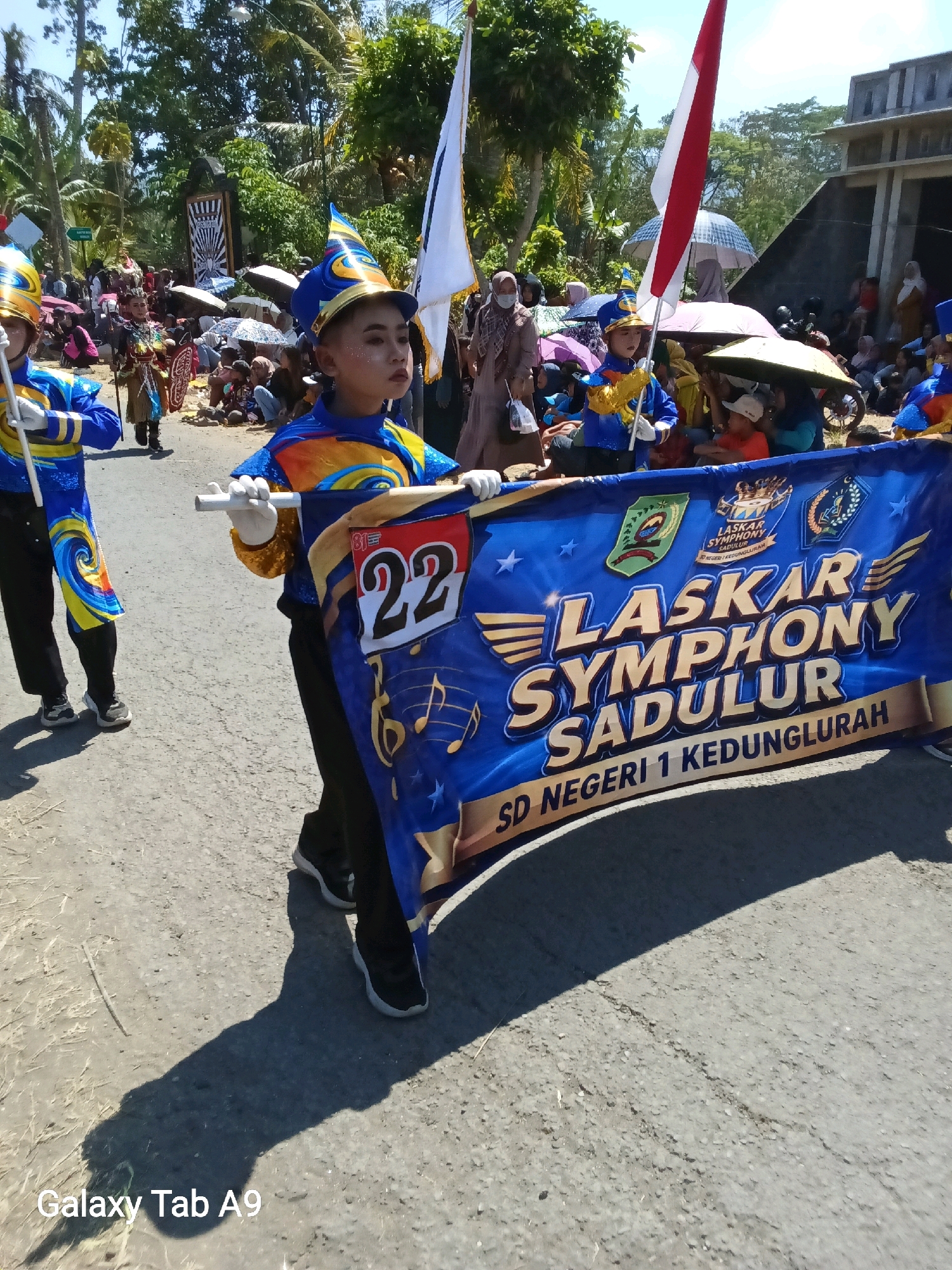 In Ngadirenggo, Indonesia, on a bright, sunny day, a group of children in marching band uniforms participate in a parade. The main focus is on a young boy in a blue and gold uniform with a distinctive feathered hat, holding a large blue banner. The banner, emblazoned with gold lettering, reads "LASKAR SYMPHONY SADULUR SD NEGERI 1 KEDUNGLURAH," with additional emblems and the number "22" prominently displayed. Another child in a similar uniform is visible to the left. The background shows a crowd of spectators, some holding umbrellas, and lush green trees lining a paved street. The architecture in the background suggests a rural or suburban Indonesian setting. The overall atmosphere is festive and celebratory.