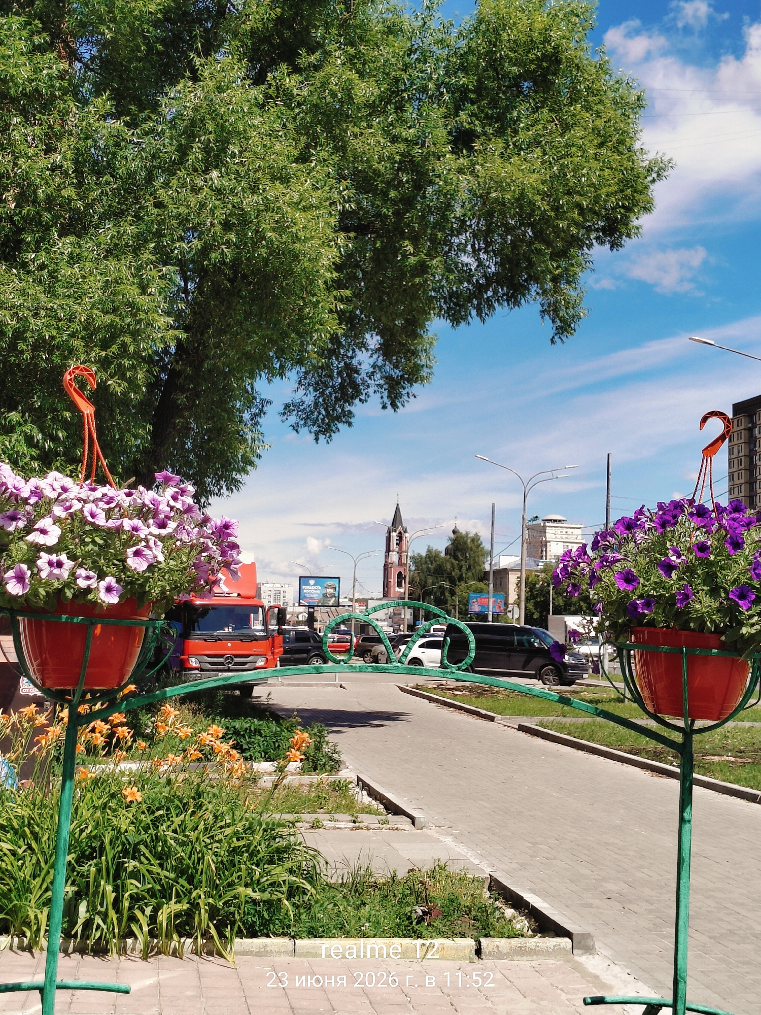 The image captures an outdoor scene on a sunny day, likely in the afternoon given the bright light and shadows. The foreground features two large, hanging pots filled with vibrant purple petunias, supported by green metal stands. Below these pots, a patch of orange daylilies adds another burst of color to the scene.
Behind the flowers, a paved walkway runs alongside a road. Several vehicles are visible on the road, including a red truck, a black van, and other cars. In the mid-ground, a red truck is prominently parked. Further in the distance, a red and black church tower with a spire stands out against the blue sky, suggesting a town or city setting. The sky is predominantly blue with scattered white clouds. A large, leafy green tree dominates the upper left portion of the image, its branches casting shadows.
The visible text at the bottom right corner reads "realme T2" and "23 июня 2026 г. в 11:52", indicating the device used to capture the image and the date and time of the photograph, which is June 23, 2026, at 11:52 AM. The overall impression is a bright, cheerful, and slightly urban environment during daytime.