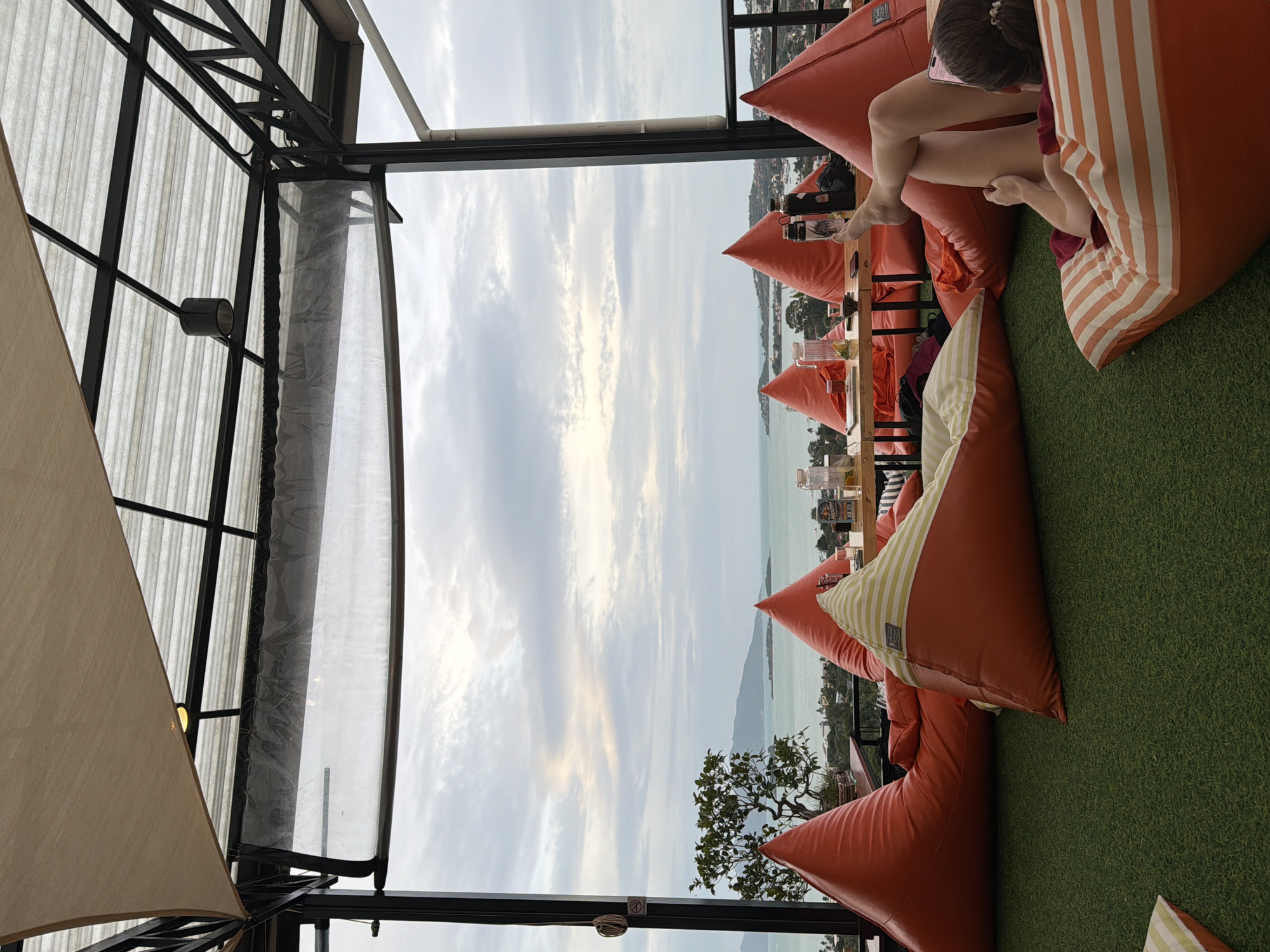 A wide-angle shot captures a rooftop seating area in Ko Samui District, Thailand, overlooking the ocean and distant islands under a cloudy sky. The foreground is dominated by several large, orange beanbag chairs scattered across a green artificial turf floor. One person is visible, reclining on a striped beanbag in the lower right, looking at a phone. A table with drinks and bottles is positioned behind some of the beanbags. The seating area is covered by a translucent roof supported by a dark metal frame.