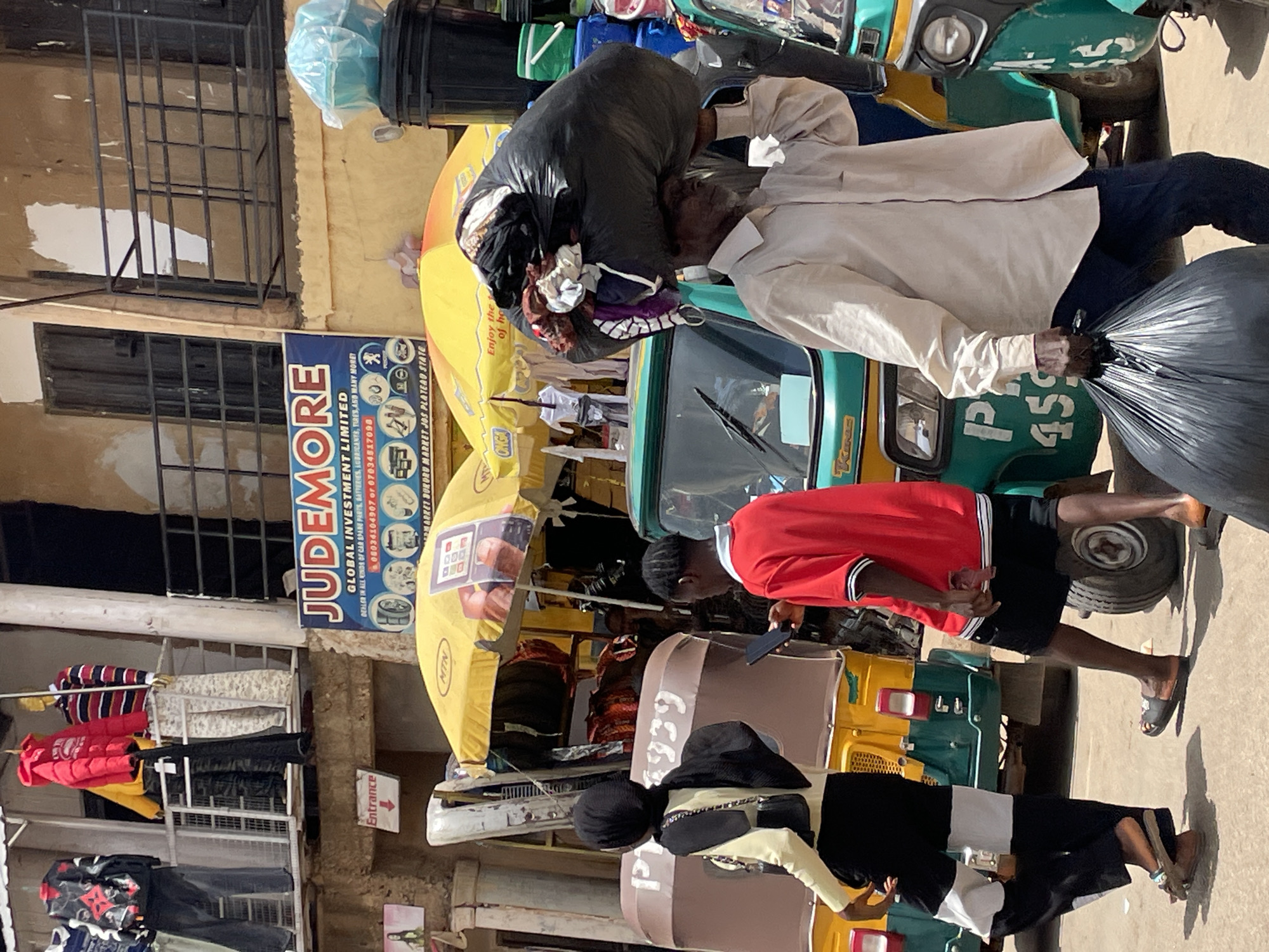 The image shows a street scene in Bukuru, Nigeria, with multiple auto-rickshaws (tuk-tuks) lined up. A sign for "JUDEMORE GLOBAL INVESTMENT LIMITED" is visible on a building; it identifies the business as a dealer in car spare parts, batteries, lubricants, tires, and other items, providing phone numbers. An MTN branded umbrella is set up near one of the tuk-tuks. People are present: one man in a light-colored shirt is carrying a large bag and another is wearing a red shirt. Clothes are displayed for sale on racks outside the building. The street surface is paved.