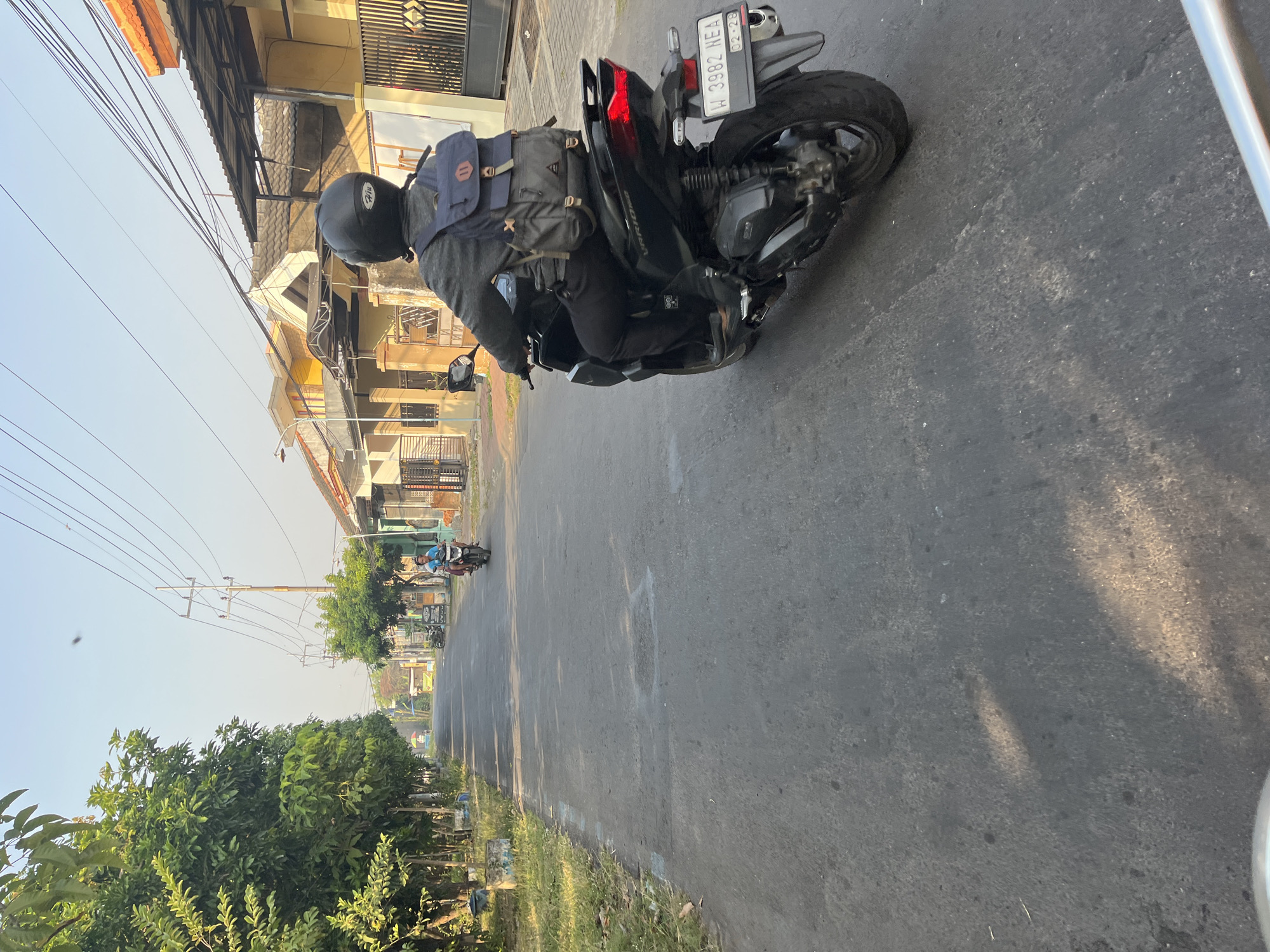 A medium shot captures a person riding a black motorcycle on a paved road in Karangtanjung, Indonesia. The rider, facing away from the camera, wears a black helmet and a gray jacket over dark pants, with a blue and gray backpack secured on their back. The motorcycle, identified by the license plate "W 3982 NEA" with an expiration date of "02-28," has a red taillight illuminated. The road is bordered by trees and grassy verges on the left, and houses with yellow and orange facades on the right. Utility poles with numerous wires are visible along the street. Another person on a motorcycle is seen further down the road in the distance. The sky is clear and light blue, suggesting daylight. The scene appears to be a residential street in a suburban or semi-urban area.