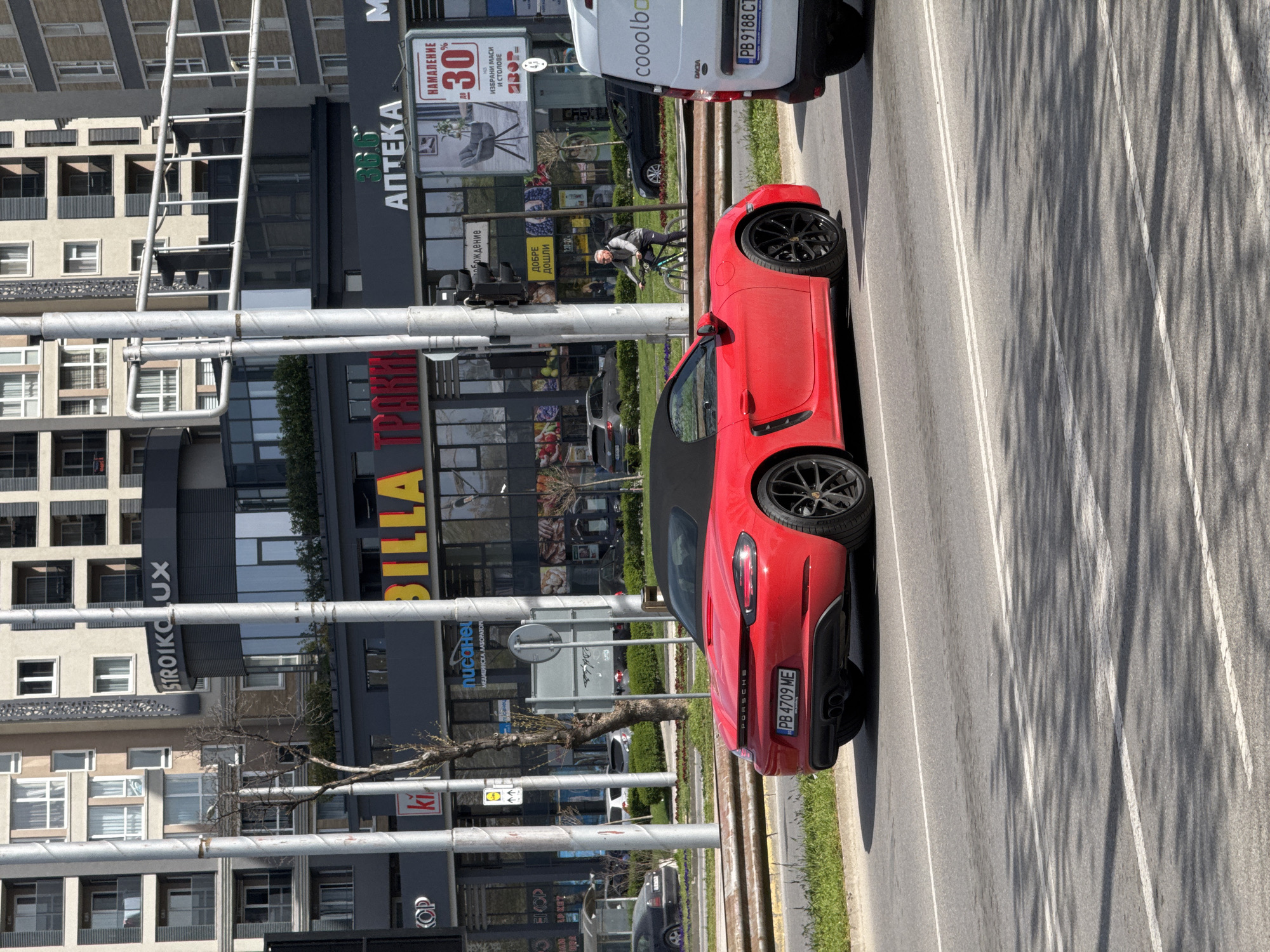 This image depicts an urban street scene in Plovdiv, Bulgaria, on a bright, sunny day. The central subject is a striking red Porsche convertible, identifiable by the "PORSCHE" emblem on its rear and the Bulgarian license plate "PB 4709 ME". It appears to be parked or slowly moving on the right side of a multi-lane road. Ahead of the Porsche, a white Dacia van with the "cooolbo" logo and license plate "PB 9188 CT" is also visible.
In the background, a row of modern multi-story buildings lines the street, housing various businesses. Visible text on these buildings includes "СТРОЙКО ЛУКС" (Stroyko Lux) and "BILLA," indicating a supermarket. Other commercial signs include "АПТЕКА" (Pharmacy) with the number "36.6," and an advertisement proclaiming "НАМАЛЕНИЕ ДО 30%" (Discount up to 30%) on "ИЗБРАНИ МАСИ И СТОЛОВЕ" (selected tables and chairs). Further signs read "ДОБРЕ ДОШЛИ" (Welcome) and "ТРАКИЯ".
A person riding a bicycle is visible in the middle ground, adding to the sense of city activity. Bare tree branches are visible, suggesting either winter or early spring. Long, distinct shadows are cast across the asphalt road, indicating a clear sky and direct sunlight. The scene portrays a typical weekday urban environment.