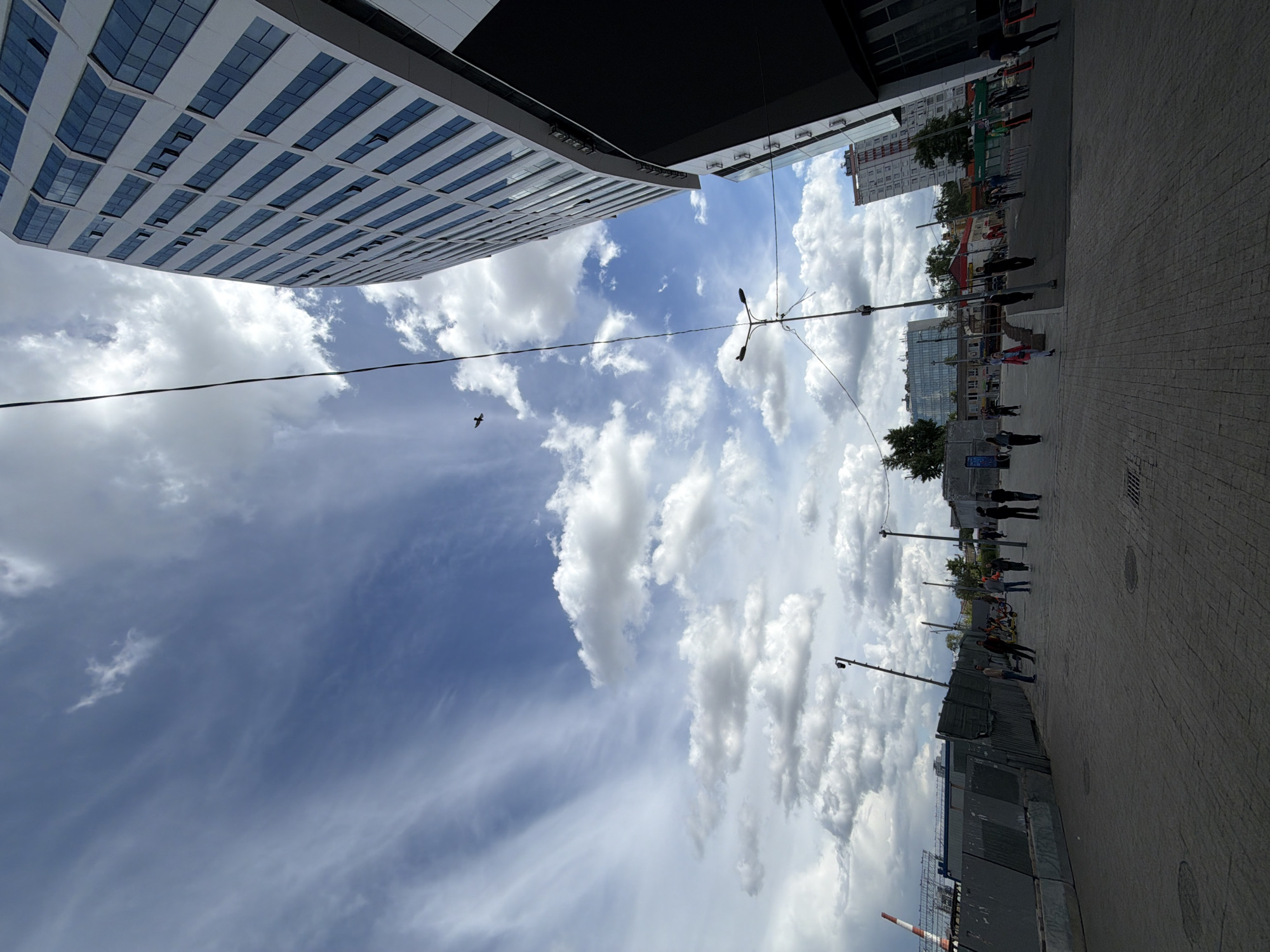 The image shows a street scene in an urban environment. A tall, modern building with a grid-like facade is visible on one side of the frame, while another building and a street lined with people and streetlights are on the other. The sky is filled with clouds. A power line is also visible, intersecting the clouds.