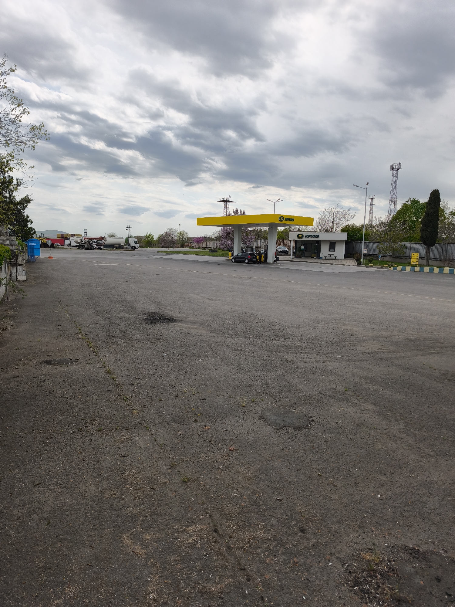 This daytime photograph depicts a gas station and large asphalt lot under an overcast sky, located in what appears to be a commercial or industrial area in Burgas, Bulgaria.
The foreground is dominated by a grey, somewhat worn asphalt surface with sparse patches of grass and weeds growing through cracks. In the midground, to the right, stands a gas station featuring a bright yellow canopy covering the fuel pumps. The canopy, along with an adjacent small white building, displays the company name "К КРУИЗ" (K KROUIZ). A black car is visible at the pumps, with a person standing next to it, likely refueling.
The background to the left features several large commercial vehicles, including white tanker trucks, parked in an open lot, suggesting a truck stop or depot. Further back, a mix of trees with green leaves, some bare trees, and trees with pink blossoms line the horizon. Tall utility poles and what seem to be communication towers are also visible in the distance.
To the far left, a blue portable toilet is present, bearing the text "ПРЕДПАЗНА ТЕХНИКА" (Protective Equipment/Safety Equipment). The gas station area is demarcated by a fence and concrete barriers painted with yellow and black stripes. The sky is uniformly covered with grey clouds, indicating an overcast day. The scene is functional and mundane, capturing a typical roadside service area.