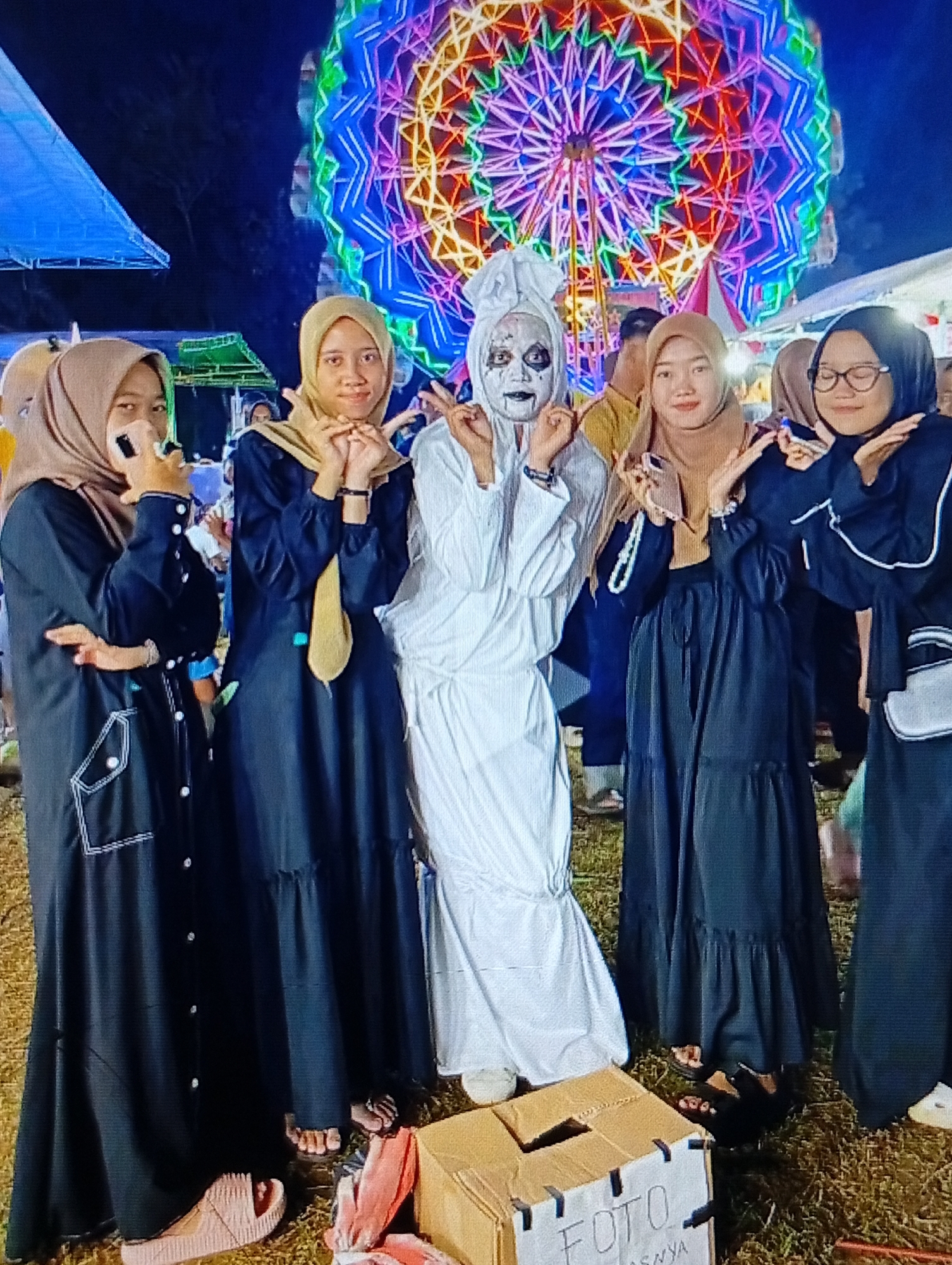 The image depicts a group of five individuals, appearing to be young women, posing for a photograph outdoors at night. In the background, a brightly lit Ferris wheel with intricate, colorful patterns is prominently displayed. To the left, a portion of a blue tent structure is visible.
The individuals are dressed in modest, long-sleeved garments. Four of them are wearing hijabs. The woman in the center is dressed in a white costume, with white face paint applied in a ghostly or theatrical manner, including dark eye makeup and dark lipstick. Her hair is covered with a white cloth, and she is striking a pose with her hands near her face. The other four individuals are wearing dark-colored dresses and hijabs of various shades of brown and beige. They are also posing, with some holding their hands near their faces in a gesture of peace or greeting.
In the foreground, a cardboard box with the word "FOTO" handwritten on it is placed on the ground. Various items of clothing, including what appears to be a pink sandal, are scattered around the box and the feet of the individuals.
The ground appears to be covered in dry grass or straw, indicating an outdoor setting at what seems to be a fair or festival. The overall atmosphere suggests a nighttime event with entertainment. While the image does not explicitly state the location, the request specifies Lhokseumawe, Indonesia, suggesting this event is taking place there.