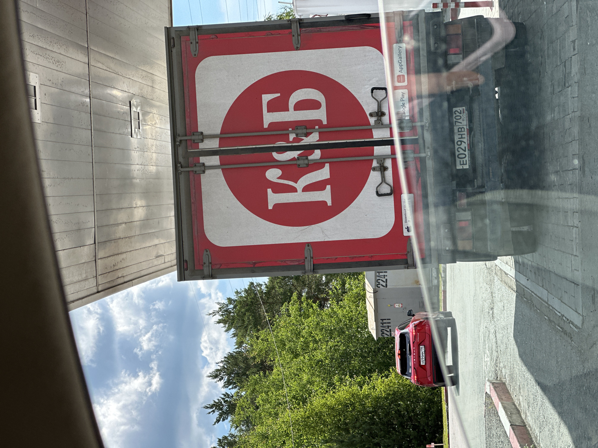 The image shows the rear of a red truck with a white circle containing the letters "К&Б" in red. The truck is parked under a metal canopy. To the left of the truck, a red SUV is visible driving away. In the background, there are trees and a cloudy sky. The license plate of the truck reads "E029HB 702". The license plate of the SUV reads "X694MM 196". A sign on the side of the truck indicates "Google Play" and "AppGallery". The scene appears to be at a gas station or a parking area. While the image itself does not provide definitive evidence of the specific location within Yekaterinburg, Russia, the presence of Russian license plates and the Cyrillic lettering on the truck suggest it is in Russia.
