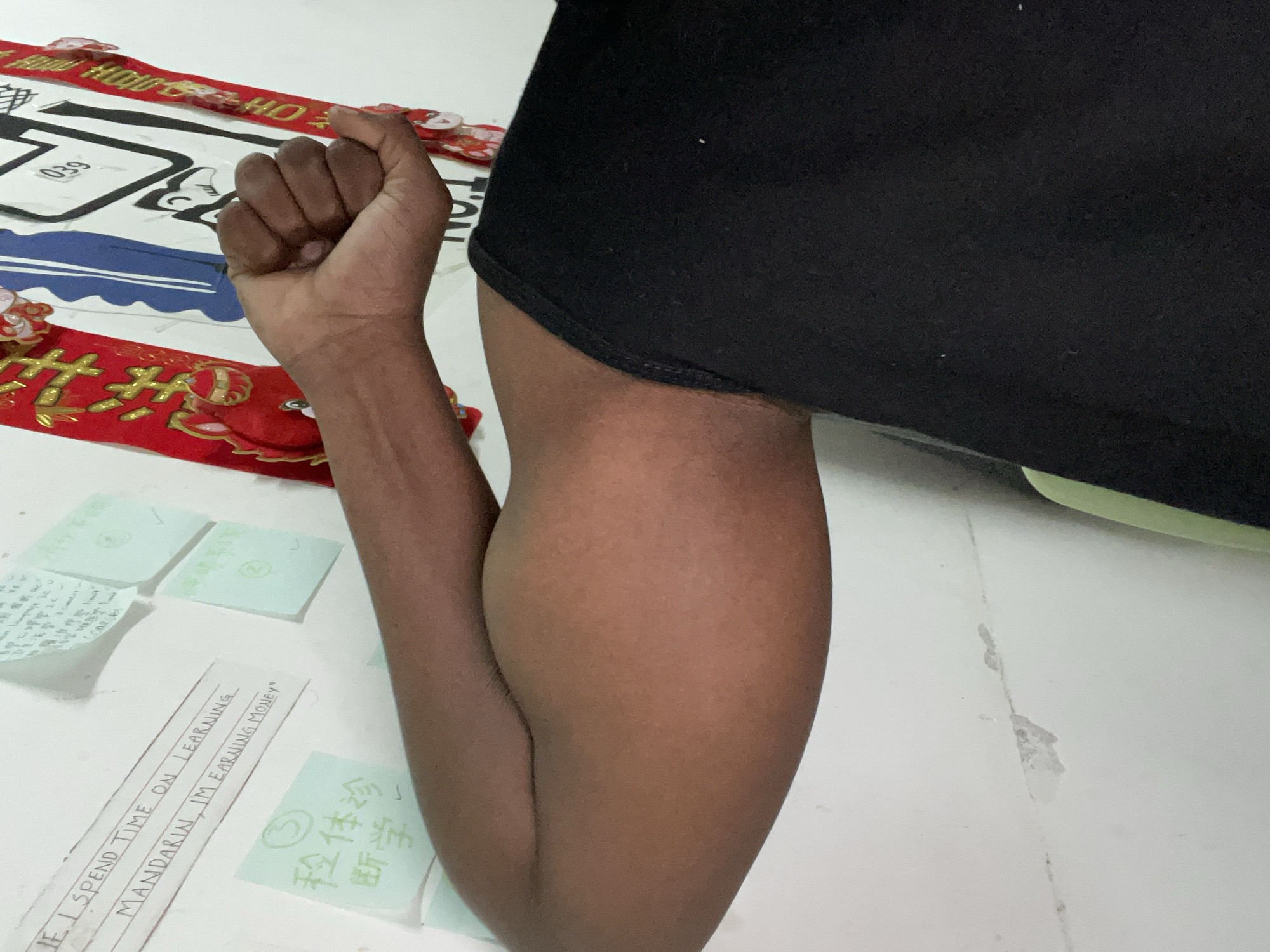 A close-up shot depicts a person's arm and fist resting on a white surface covered with papers and decorative items. The person is wearing a black garment, likely a t-shirt. The papers contain Mandarin script and English text. One paper reads "IF I SPEND TIME ON LEARNING MANDARIN, I'M EARNING MONEY". There are also decorative red and gold Chinese New Year-themed banners visible. The scene is likely in Chongqing High-tech Zone, China, based on the presence of Mandarin text.