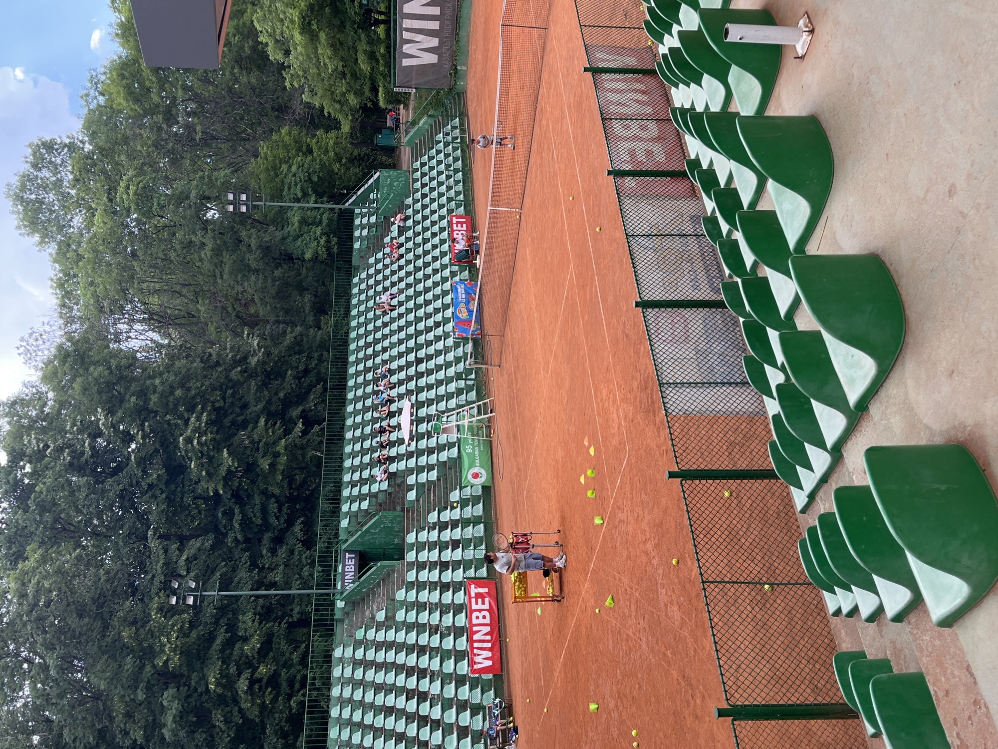 This media file captures a tennis court with clay surface and green spectator seating. The scene is outdoors, under what appears to be daytime conditions, with a bright sky and scattered clouds visible amongst the surrounding lush green trees. The court is set up for practice or a match, with tennis balls scattered on the surface and some placed near a practice machine.
Several people are visible. On the court, a person in a white shirt and shorts is engaged in a tennis activity, possibly practicing or participating in a drill. Another individual is standing near a tennis ball hopper and a practice machine. In the spectator stands, a few people are seated, observing the court.
Notable details include the vibrant green of the stadium seats, which are arranged in multiple tiers. A net divides the tennis court. Several banners with text are visible. One banner prominently displays "WINBET" in white letters on a dark background. Another banner also features "WINBET" and "95 years BULGARIAN TENNIS FEDERATION". The overall atmosphere seems focused on the sport, with a sense of ongoing activity. The presence of trees suggests a natural or park-like setting for the tennis facility.
