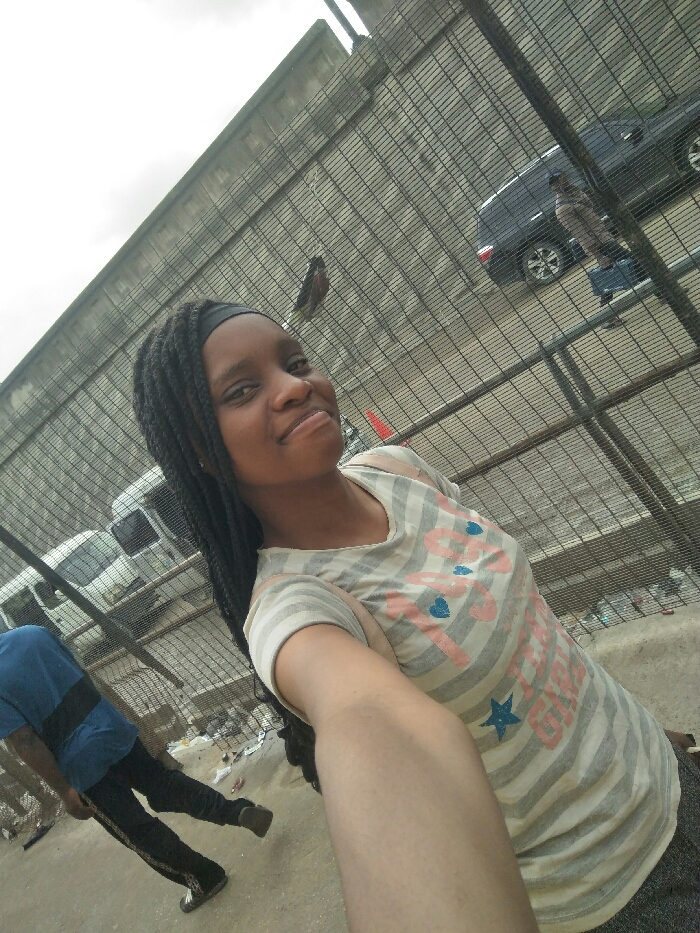 A young female, identified as the primary subject, is taking a selfie outdoors in Ajah, Nigeria. She is positioned in the foreground, slightly angled, extending her right arm towards the camera. She has a dark complexion, wears a black headband, and her long, dark braided hair falls over her left shoulder. She is dressed in a light-colored, possibly off-white or light grey, horizontally striped t-shirt with "1997" printed in pink, along with blue heart and star motifs. Her expression is a faint smile, with her eyes directed at the camera.
A dark metal grid fence separates the foreground from the background street scene. Through the fence, a paved road is visible, displaying varying amounts of litter and a small orange traffic cone near the center-right. On the road, a dark-colored SUV is visible to the right, and a white van is present to the left. Two individuals are also visible in the background street area: one male figure, partially obscured on the left, wearing a blue t-shirt with a horizontal black stripe and dark trousers, walking away from the camera. Another individual is faintly visible on the right, behind the dark SUV, wearing light-colored upper clothing and blue lower clothing. The sky is overcast, and a concrete structure is visible in the upper background, beyond the fence.