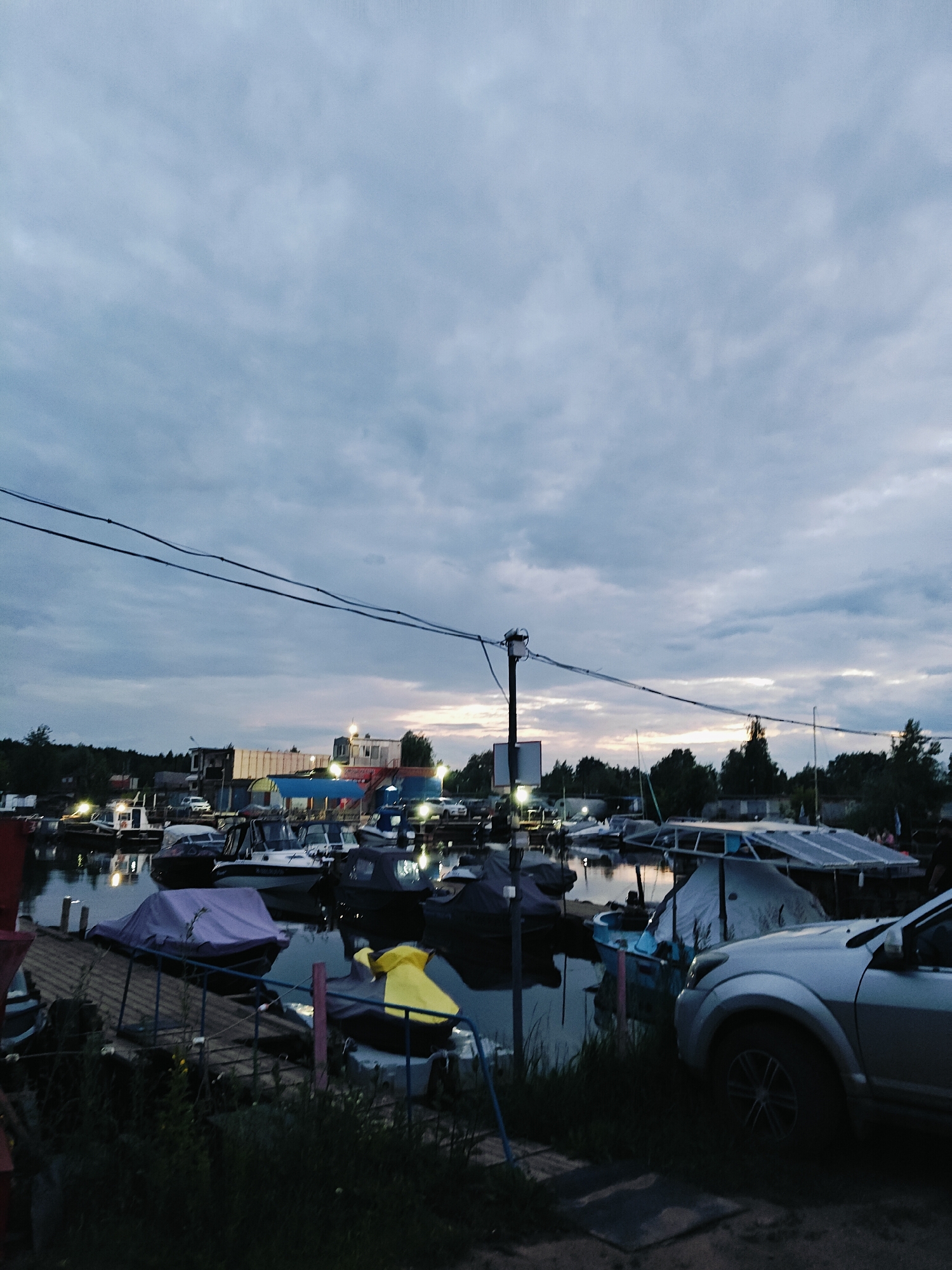 The image depicts a dusk scene at a marina in Chaikovsky, Russia, likely on the Kama River. The sky is overcast with a hint of sunset color peeking through the clouds. Numerous boats are moored in the water, many covered with tarps of various colors, including purple and grey. Some boats are smaller motorboats, while others appear to be larger yachts or houseboats. In the background, several structures are visible, including what appear to be small buildings, possibly workshops or residences, with lights illuminating them. A white SUV is parked in the foreground on the right side of the image, partially obscuring the view of some boats. Power lines are strung across the upper part of the frame. The overall impression is one of a functional, somewhat rustic, waterfront community.